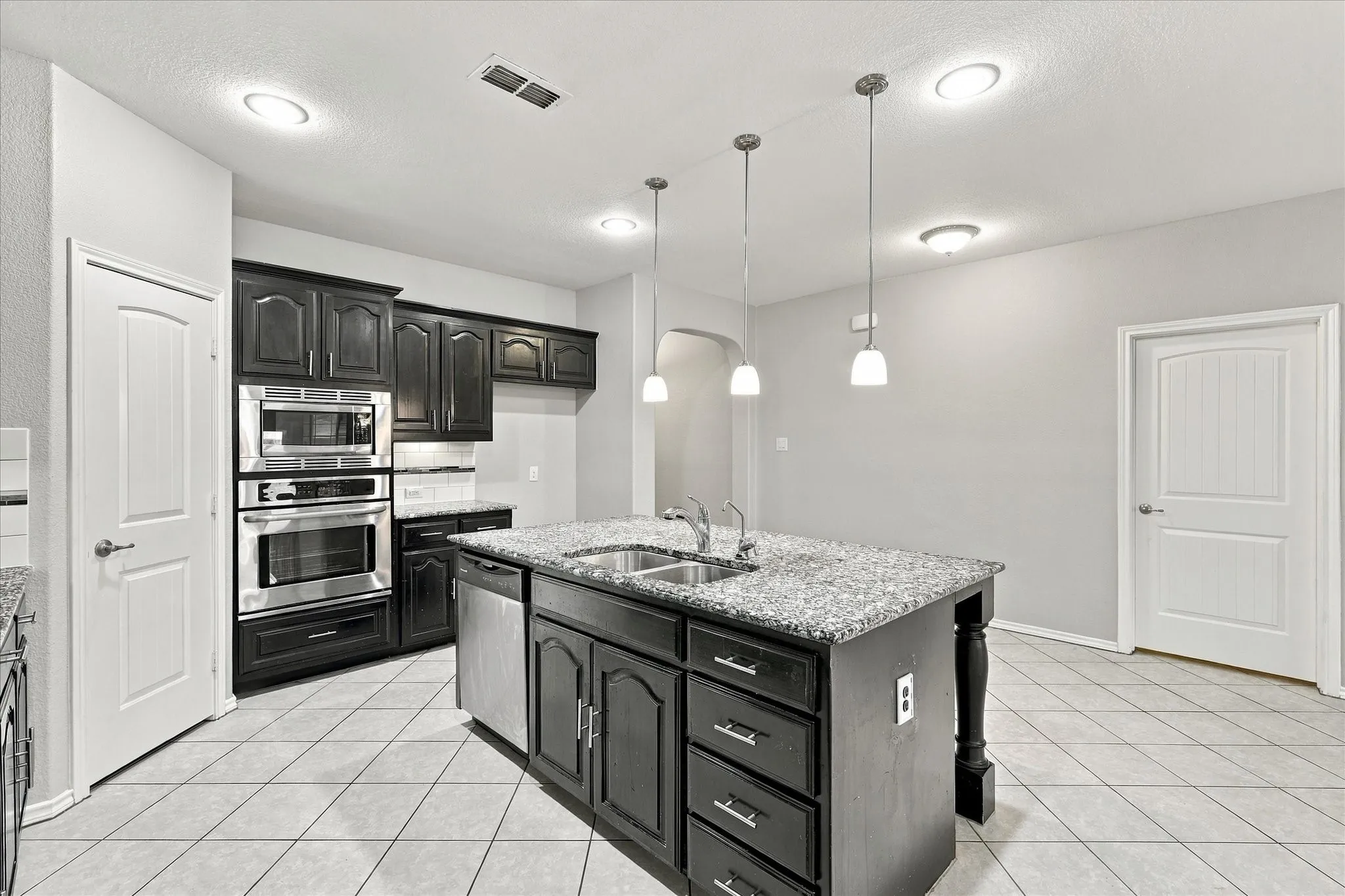 Kitchen featuring dark wood cabinetry, stainless steel built-in appliances, a central island with a sink and granite-style countertop, and white tile flooring