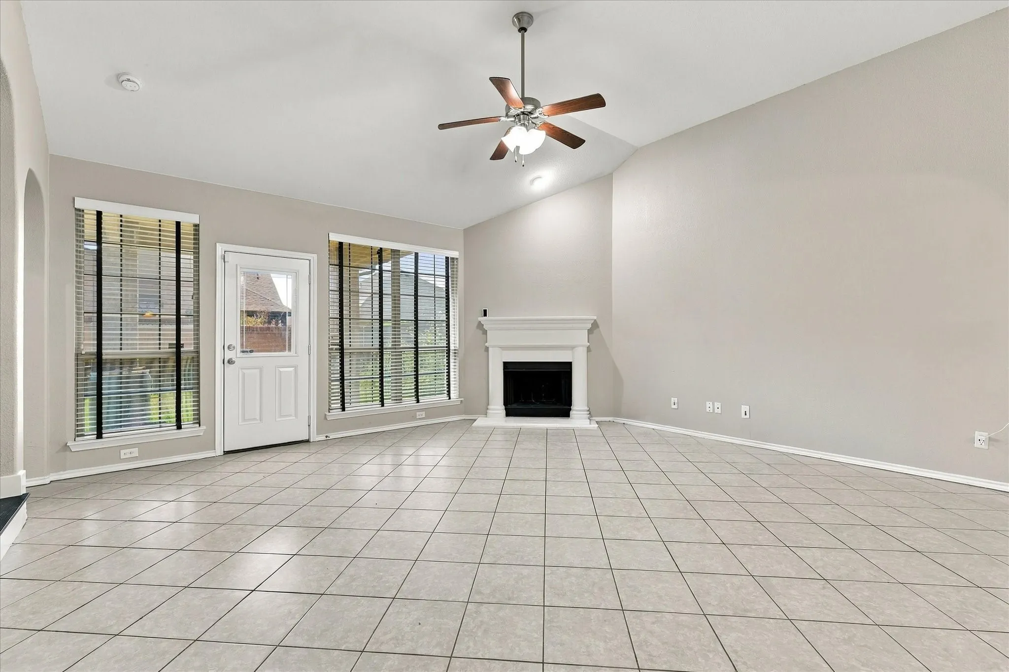 Vaulted ceiling, ceramic tile flooring, ceiling fan with light fixture, white fireplace with mantel, and direct outdoor access