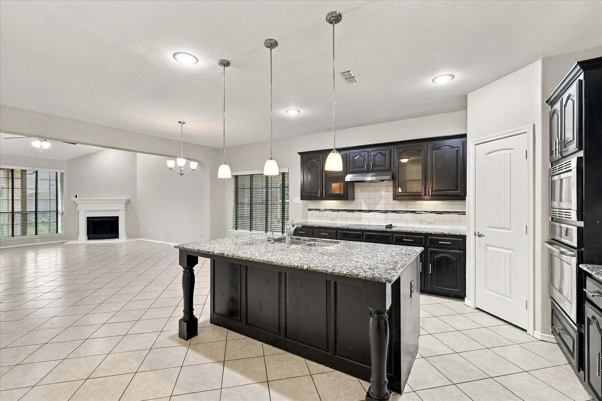Kitchen island with granite countertop and pendant lighting, dark wood cabinetry, tile flooring, built-in stainless steel appliances, and a white mantel fireplace in the adjacent room
