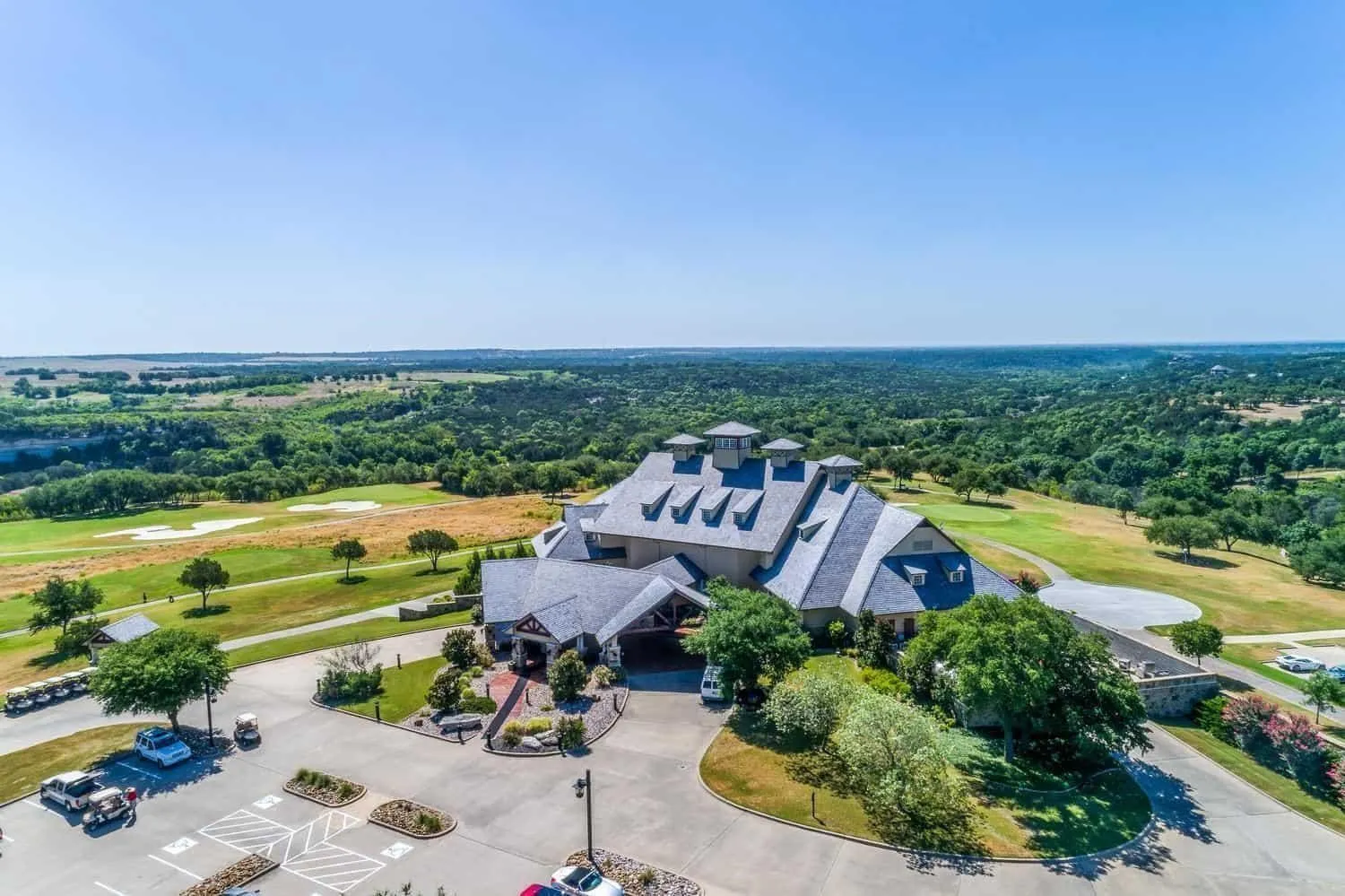 Expansive property featuring a multi-level building with a pitched roof, surrounding golf course, and a large paved parking area
