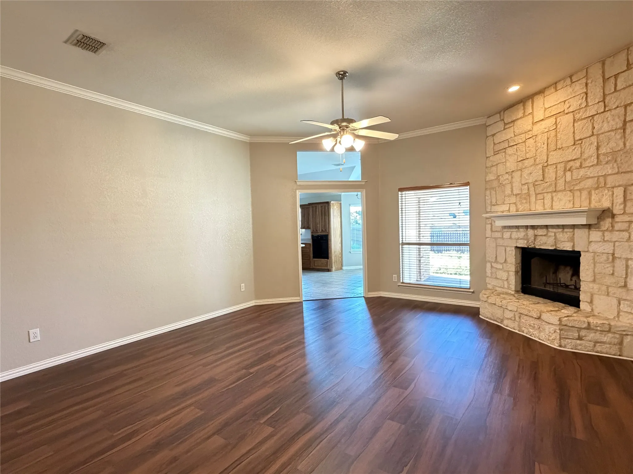 Spacious room featuring wood-finish flooring, a stone-clad fireplace with a mantel, crown molding, and a ceiling fan