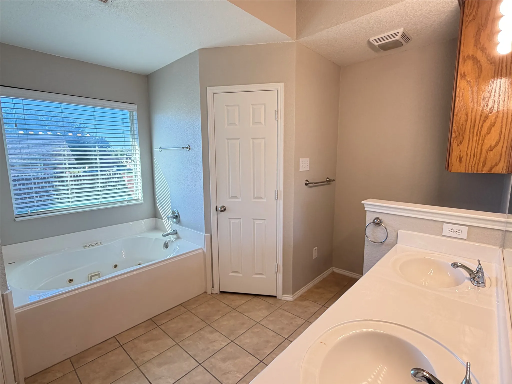 Jacuzzi bathtub with a window, dual vanity sinks, ceramic tile flooring, and a wood-finish wall-mounted cabinet