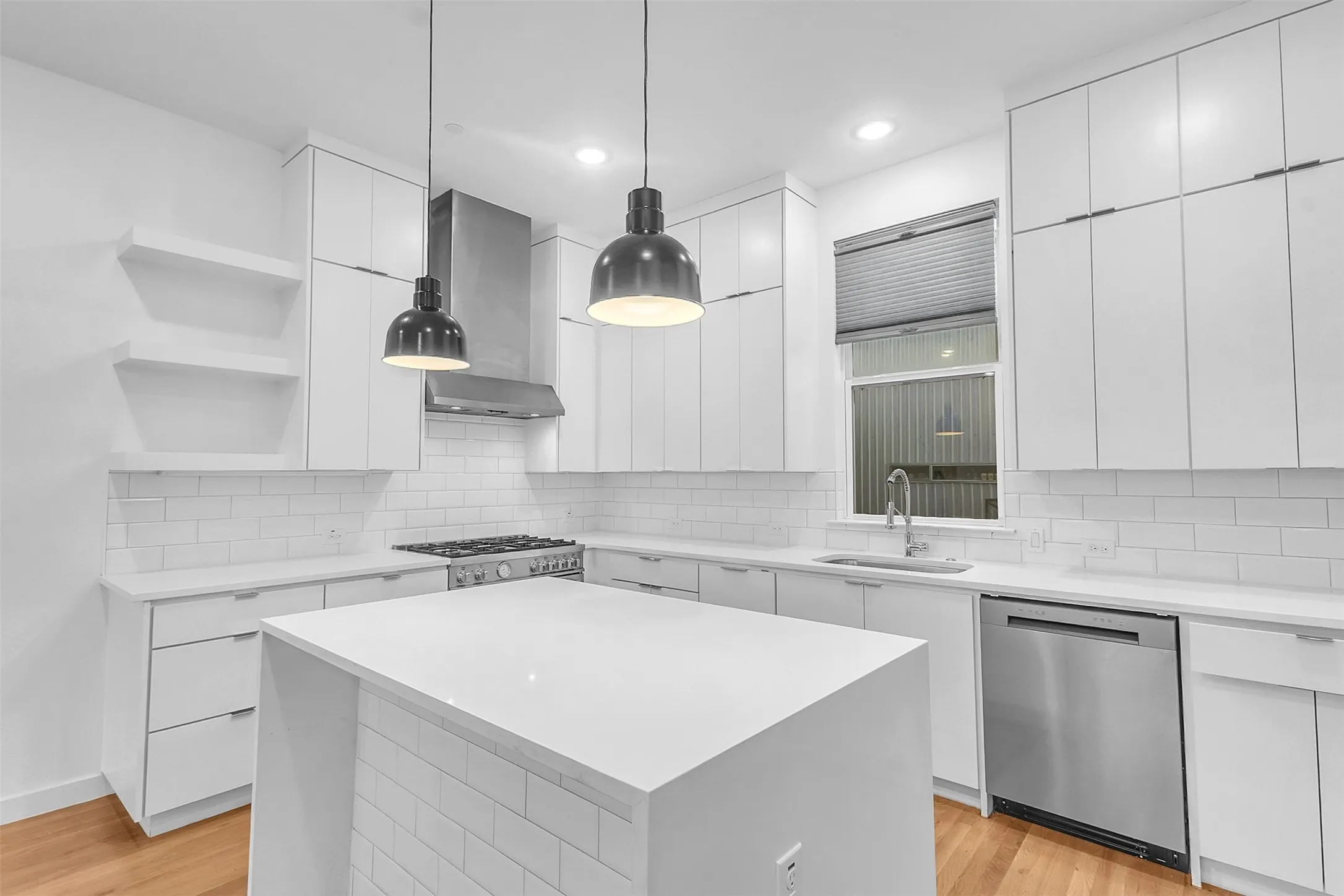 White kitchen with a waterfall island, stainless dishwasher, gas range, pendant lights and full-height cabinetry