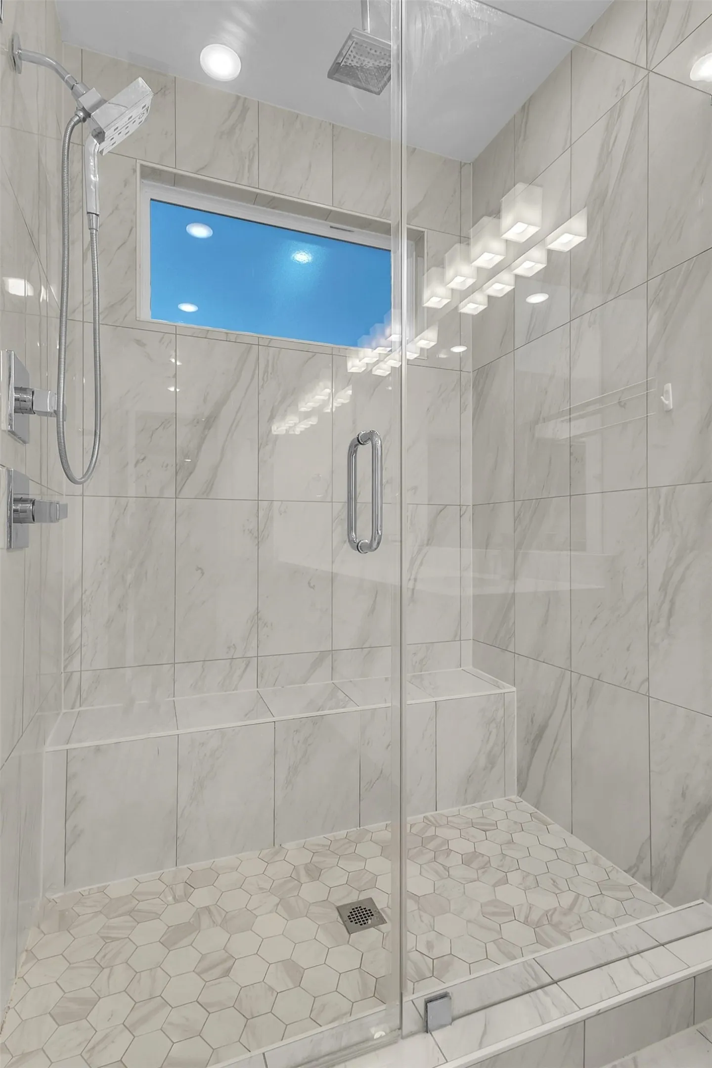 Primary walk-in shower with a frameless glass door, built-in bench, rain head, handheld sprayer and hex tile floor