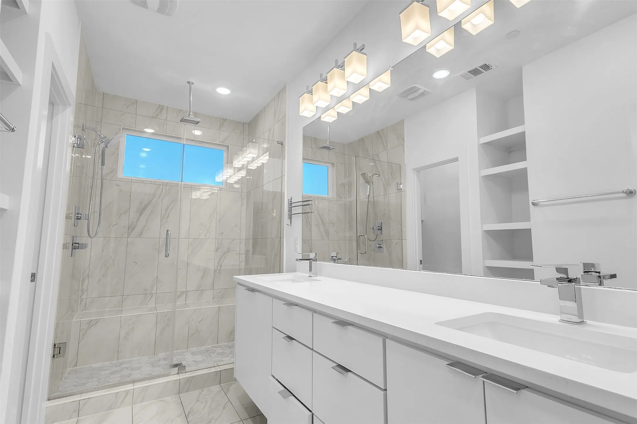 Primary bath with a glass-enclosed walk-in shower, dual-sink white vanity, wide mirror and marble-look tile