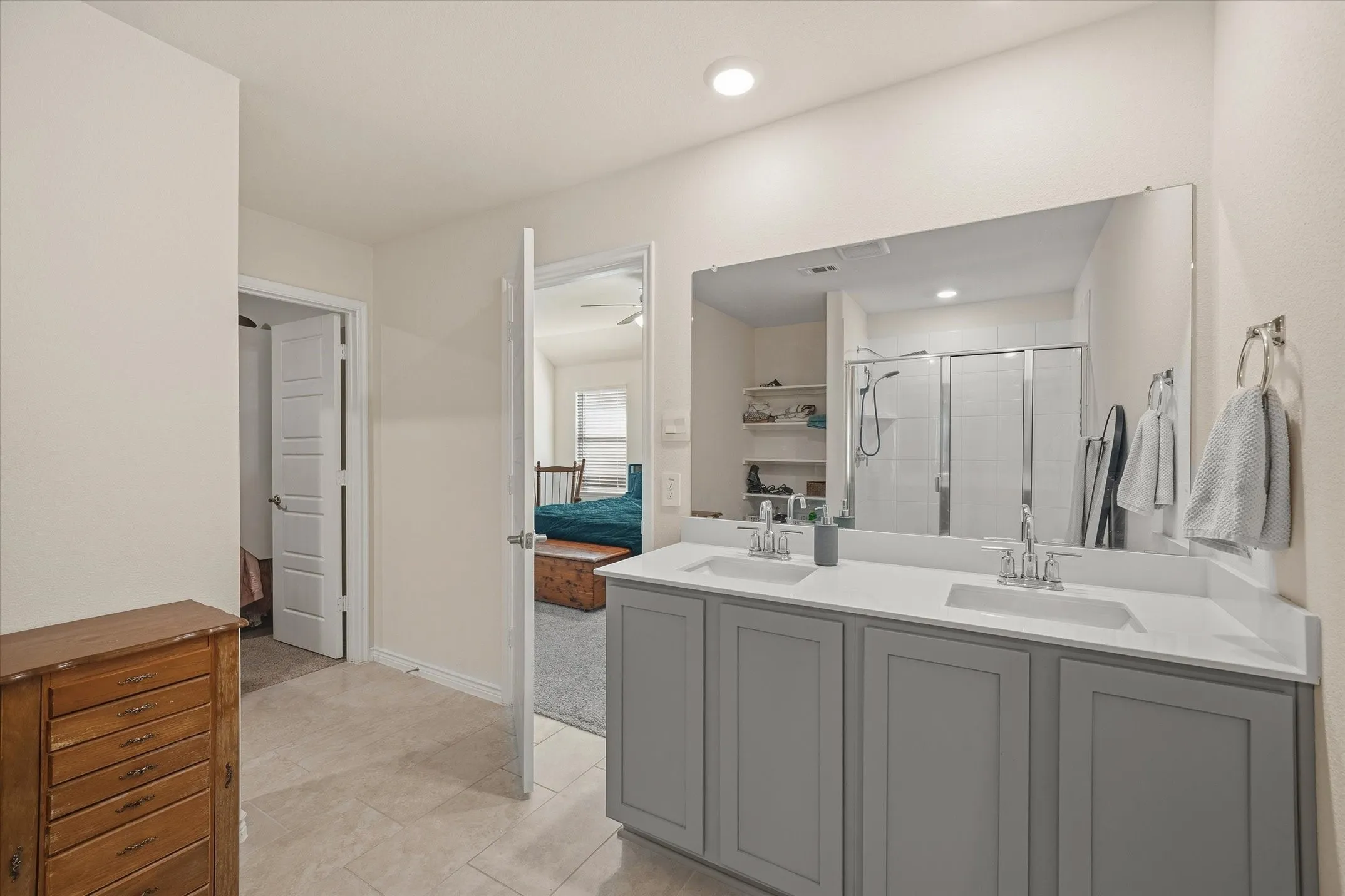 The spacious primary en-suite bathroom at 202 Buckboard Ln features an expansive dual-sink vanity with crisp white countertops, undermount basins, and slate gray shaker cabinetry. Neutral ceramic tile flooring extends throughout the space, complemented by recessed lighting and a wide frameless vanity mirror reflecting the glass-enclosed walk-in shower with vertical tile surround. Open doorways provide seamless transitions to a secondary closet area and the carpeted primary bedroom, which showcases a pitched ceiling, modern ceiling fan, and abundant natural light in this refined Caddo Mills home.
