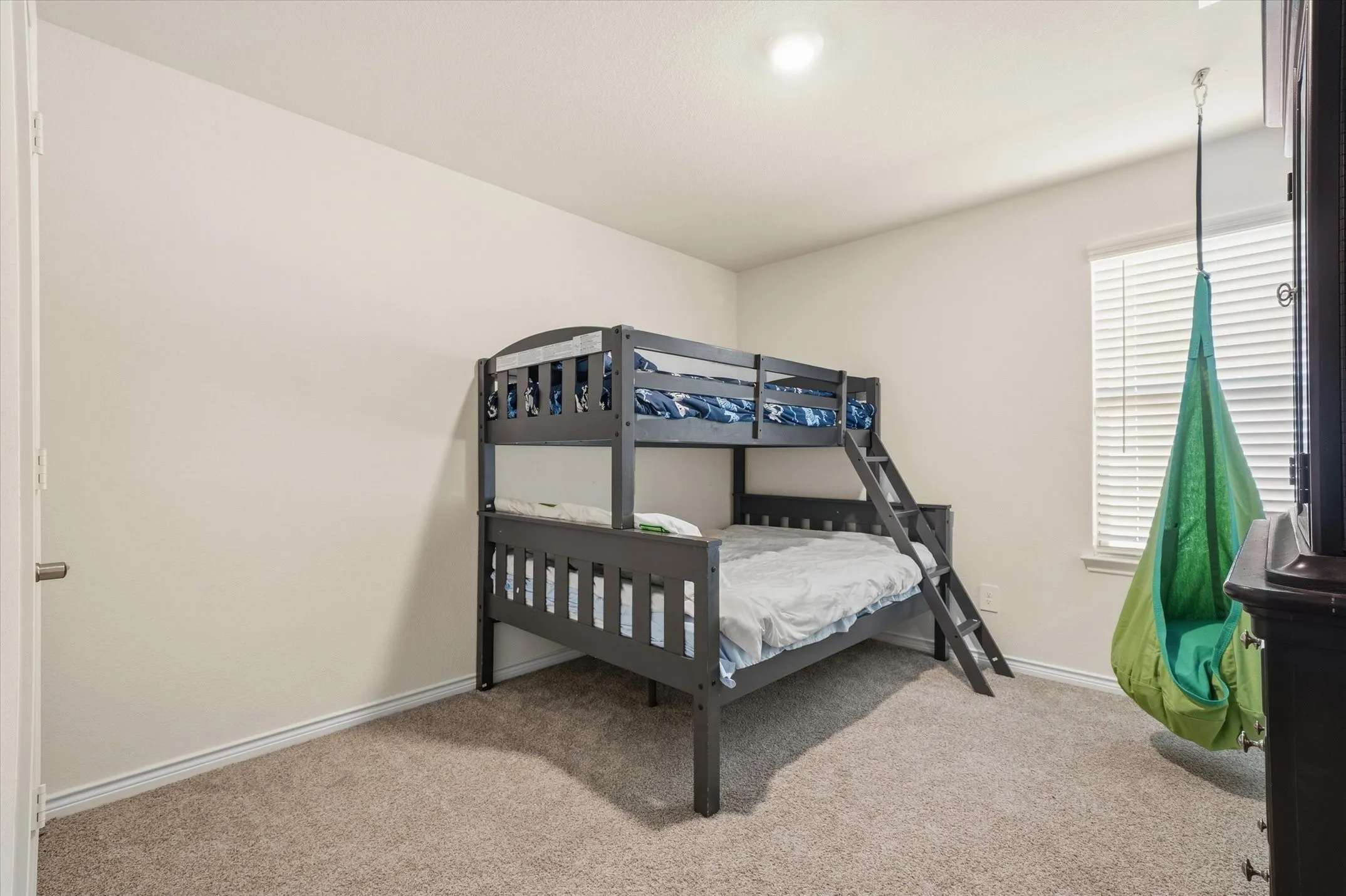 Sunlight filters through a single-hung window dressed with horizontal blinds, illuminating a comfortable secondary bedroom at 202 Buckboard Ln. The functional layout features soft neutral walls, crisp white baseboards, and durable low-pile carpeting throughout. A dark gray twin-over-full bunk bed makes efficient use of the corner, while generous floor space accommodates a playful suspended pod swing and complementary bedroom furnishings. Positioned near a white interior panel door and bathed in flush-mount ceiling light, this adaptable bedroom offers practical comfort within this inviting Caddo Mills residence.
