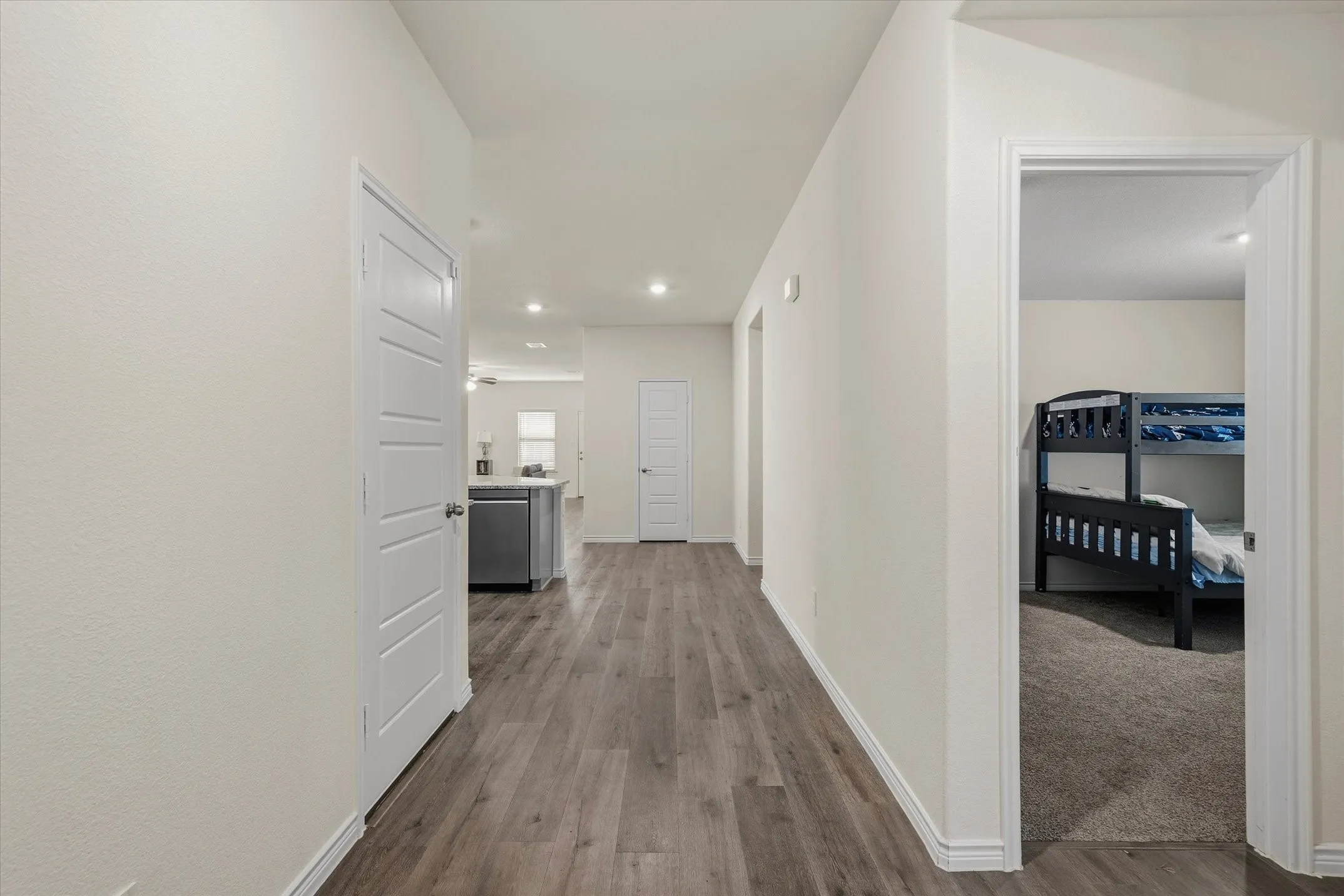 Inside this home at 202 Buckboard Ln, a wide central hallway showcases durable, cool-toned wood-grain plank flooring paired with crisp white trim. Recessed LED lighting illuminates the path as it transitions seamlessly past a carpeted secondary bedroom on the right, which comfortably accommodates bunk bedding. Ahead, the corridor opens into an airy open-concept living space and kitchen centered around a stone-topped island with a stainless steel dishwasher, highlighting a thoughtfully designed single-story interior layout crafted for modern, functional everyday living.
