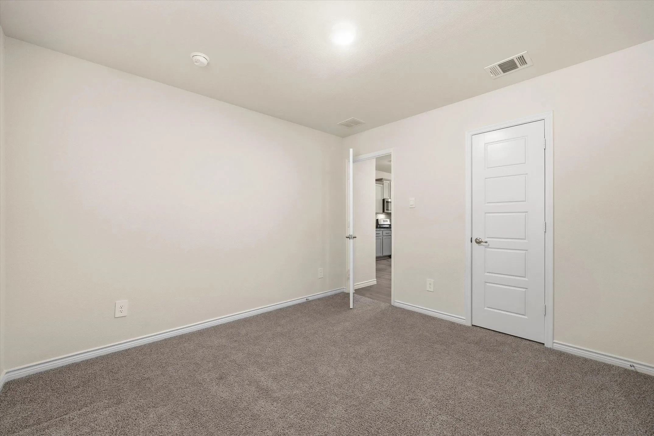 Move-in ready secondary bedroom at 202 Buckboard Ln in Caddo Mills features plush heather-gray carpeting, crisp white baseboards, and neutral walls ready for custom styling. The clean ceiling includes integrated overhead lighting and dedicated HVAC vents. Two classic five-panel interior doors with brushed metal hardware provide dedicated closet space and direct hallway flow, offering a sightline into the home's open-concept kitchen with luxury vinyl plank flooring and contemporary shaker cabinets. This versatile, well-appointed flex space delivers ideal comfort for a guest room or private home office.