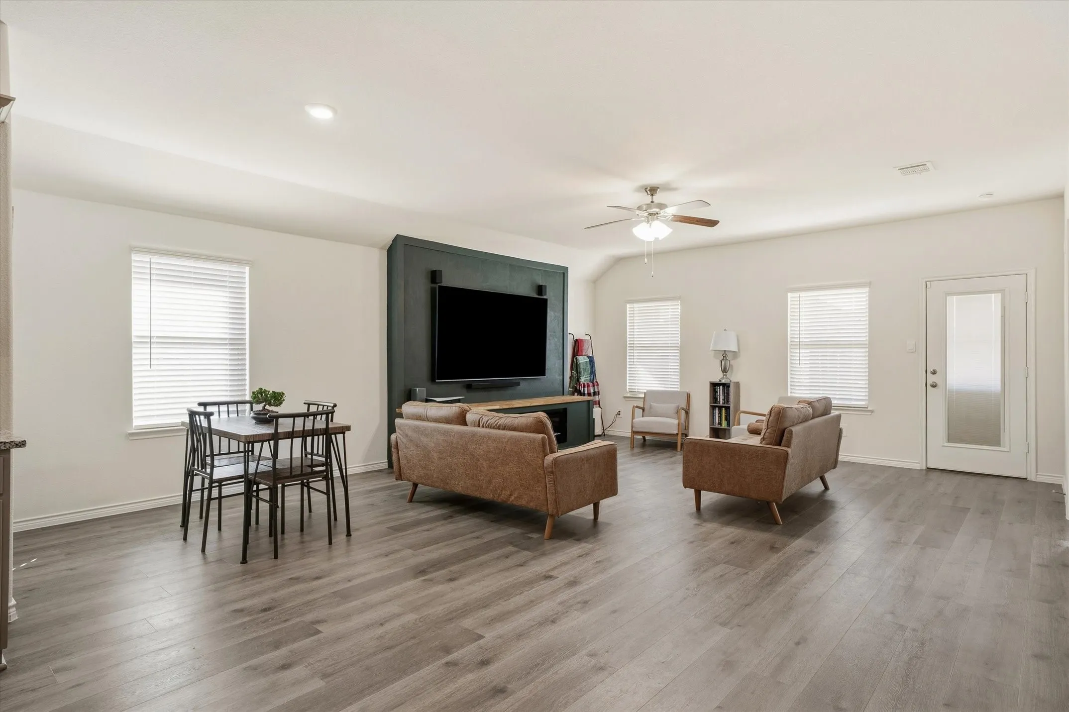 The spacious open-concept living area at 202 Buckboard Ln offers a fluid, modern layout connecting the primary family room, dining space, and adjacent kitchen. Durable wood-look plank flooring extends beneath high ceilings equipped with recessed lighting and a classic ceiling fan. A deep hunter green accent wall serves as a central media focal point, while multiple single-hung windows fitted with blinds introduce soft natural light throughout. Direct exterior access via a frosted-glass door provides a seamless transition to the backyard, perfect for effortless indoor-outdoor Texas entertaining.