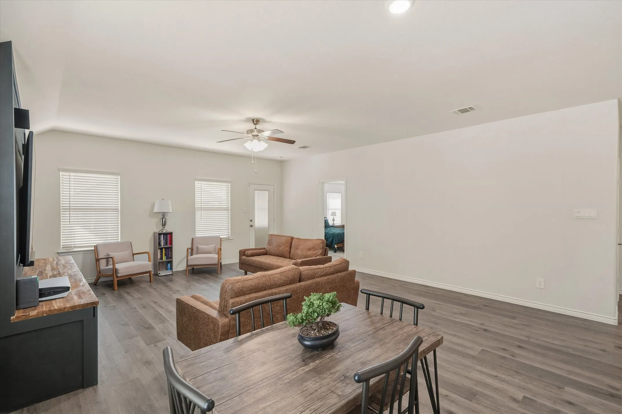 The main living area inside 202 Buckboard Ln showcases a bright, open-concept floor plan designed for effortless daily living. Durable faux-wood luxury vinyl plank flooring runs beneath neutral off-white walls, seamlessly unifying the dining space and living room. Double-hung windows with faux-wood blinds and a glass-inset exterior door along the back wall fill the interior with daylight. Modern recessed lights and a ceiling fan complete the single-story layout, which features clear sightlines toward an adjoining bedroom wing for practical everyday convenience.