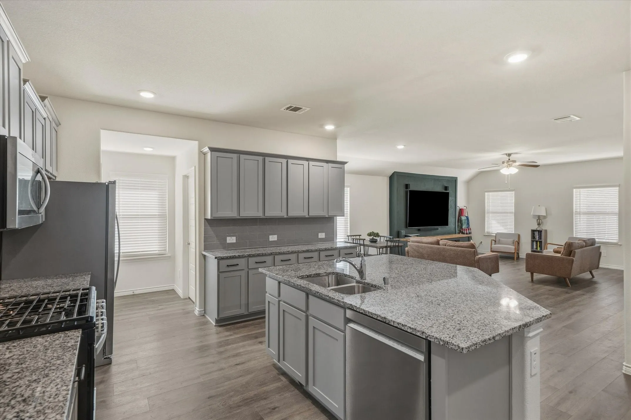 Interior view at 202 Buckboard Ln shows a contemporary open-concept layout centered around an expansive angled kitchen island. Speckled granite countertops, an undermount double-basin sink, and grey Shaker-style cabinetry accent the cooking space, which features a gas range and stainless steel appliances. Continuous wood-look luxury vinyl plank flooring unites the kitchen, bright dining alcove, and connected living room, where natural light streams through multiple blinded windows toward a tailored dark teal entertainment wall. Recessed ceiling lights illuminate this functional Caddo Mills home interior.