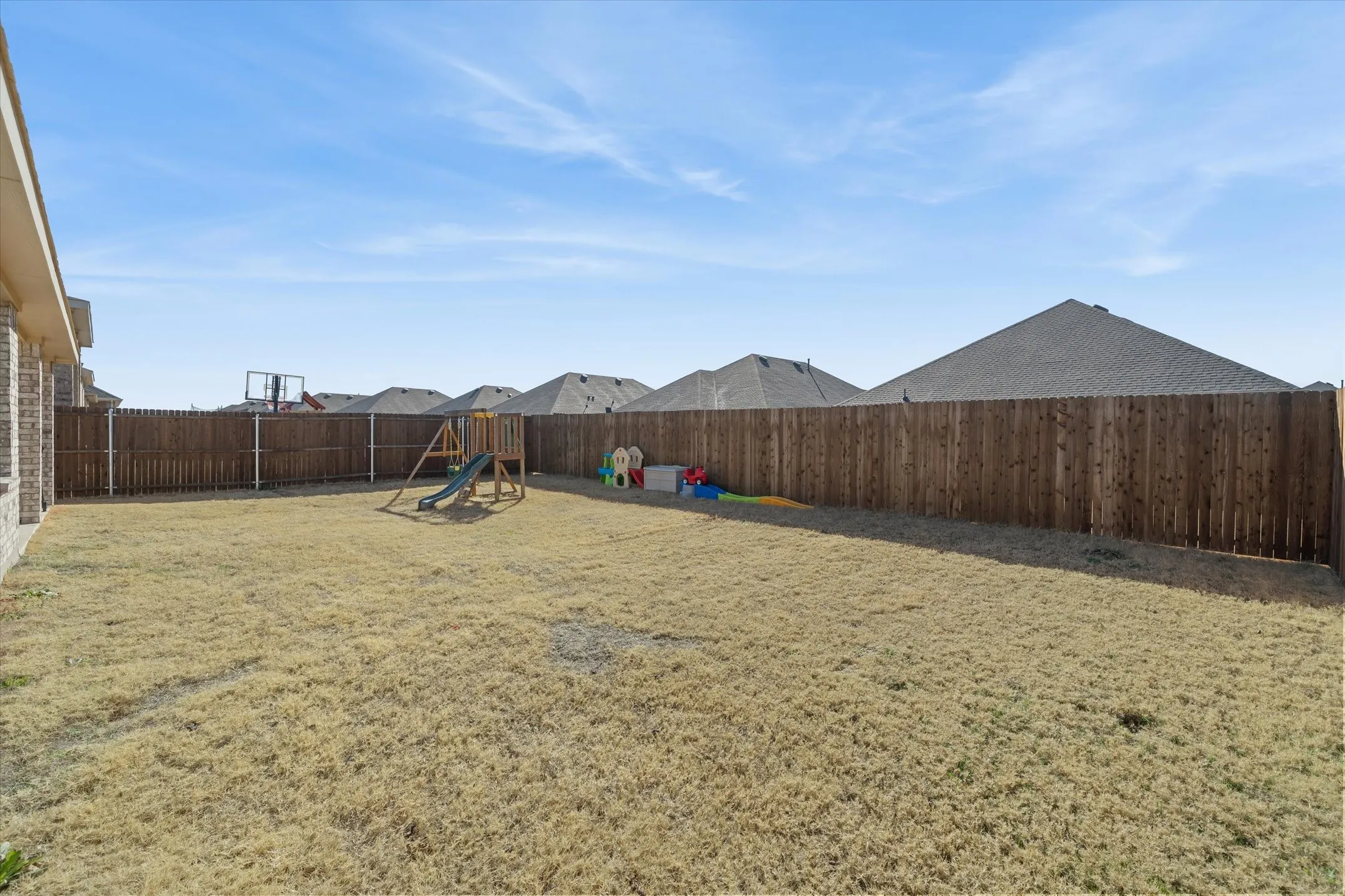 Expansive fully fenced backyard at 202 Buckboard Ln in Caddo Mills, offering a private outdoor retreat within a welcoming residential subdivision. The level yard is completely secured by a tall wood privacy fence with metal reinforcement posts, bordering neighbor single-family homes beyond. Ideal for play and recreation, the open lawn comfortably accommodates outdoor playsets and toys alongside the brick exterior of the primary home. Bright daytime sunlight highlights the generous outdoor footprint, presenting boundless potential for custom landscaping, pets, entertaining, and outdoor living.