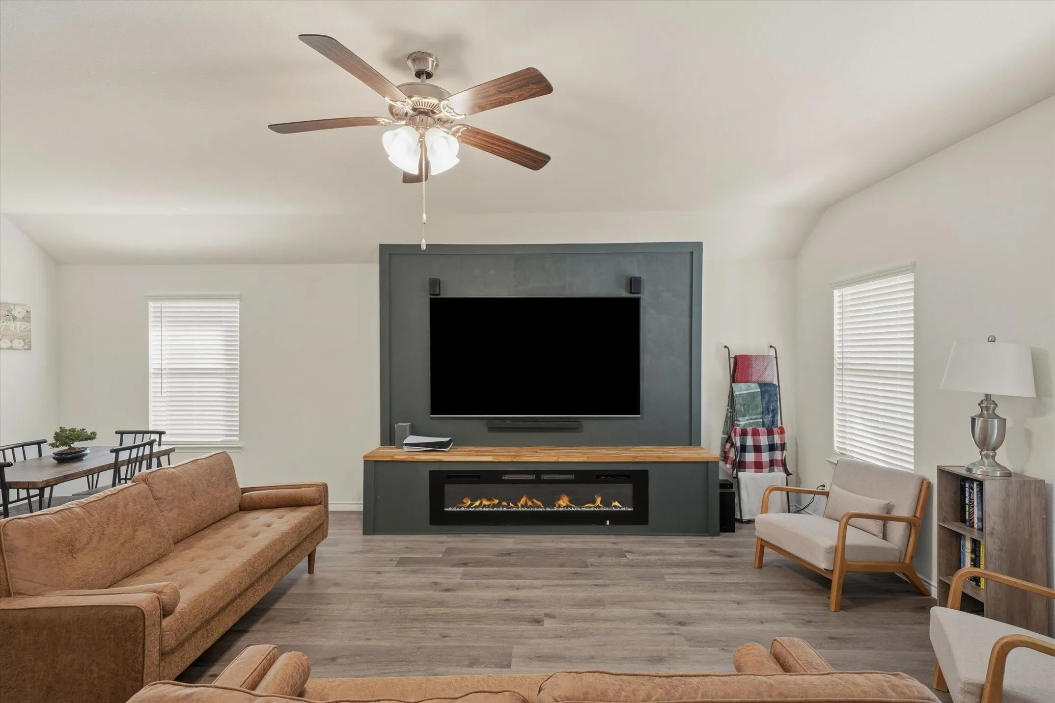 The spacious living room at 202 Buckboard Ln showcases an inviting open-concept layout centered around a custom dark-teal media accent wall. A linear electric fireplace set beneath a natural wood mantel serves as a cozy focal point below the wall-mounted television. Natural light filters through dual windows flank the fireplace, illuminating wide-plank wood-look flooring and soaring vaulted ceilings equipped with a modern ceiling fan. This comfortable interior effortlessly connects living and dining spaces, offering a warm and stylish everyday gathering environment.