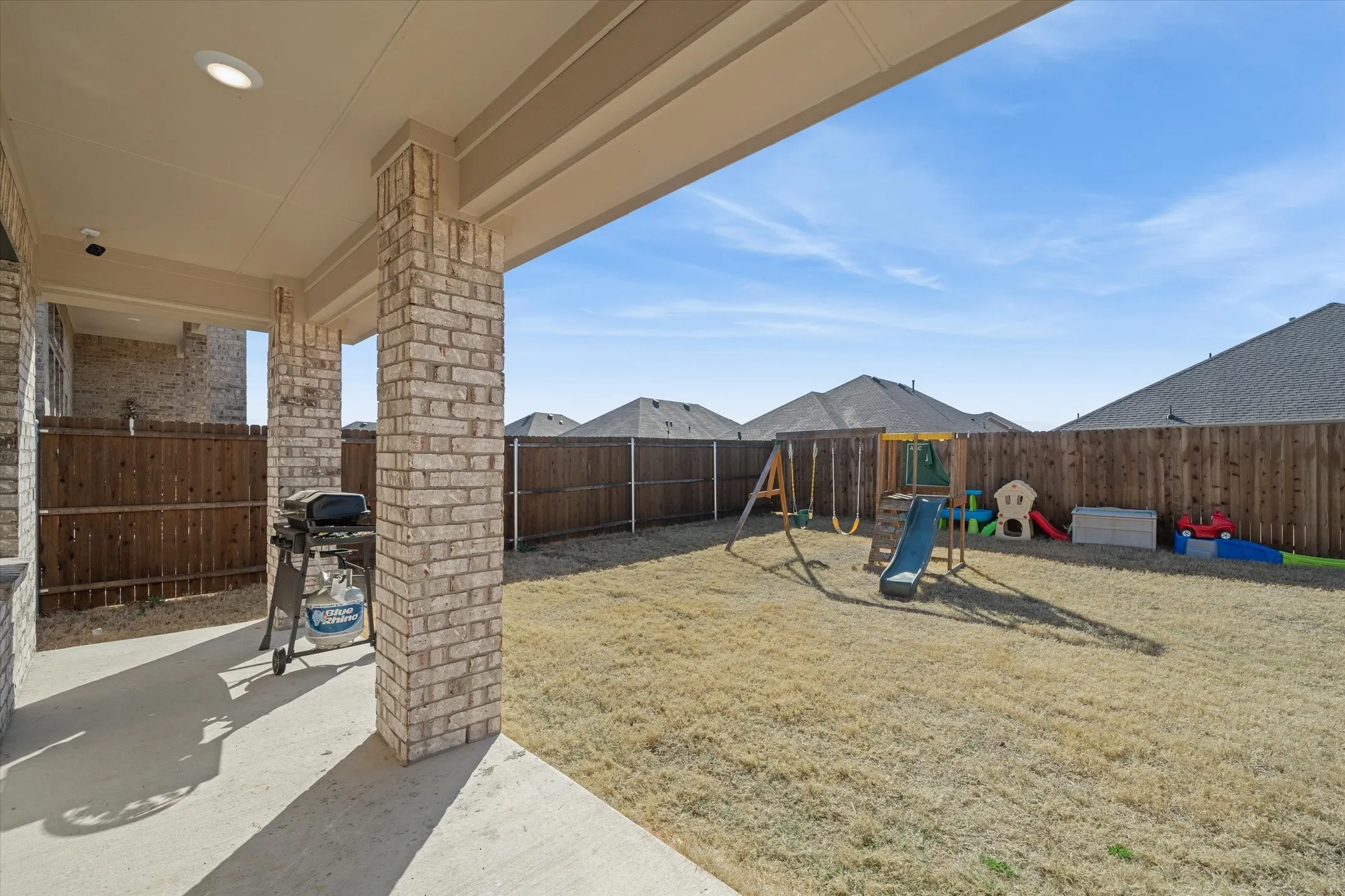 A shaded concrete back patio supported by handsome brick masonry columns offers an inviting outdoor retreat at 202 Buckboard Ln. The expansive, flat backyard is fully secured by full-height wood privacy fencing with durable metal posts, creating a private outdoor living space ideal for pets, recreation, and open-air entertaining. Recessed lighting under the covered porch accommodates evening gatherings, while the level lawn provides plenty of room for play structures or future landscape design beneath open Texas skies in Caddo Mills.