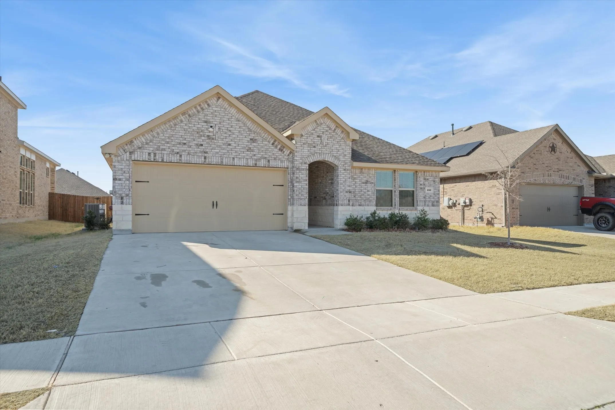 Located at 202 Buckboard Lane in Caddo Mills, this single-story suburban residence presents strong curb appeal within a master-planned community. The front elevation showcases an attractive multi-tone brick and limestone facade beneath multi-gabled asphalt shingle roofing. A spacious double-car garage with carriage-style accents anchors the wide concrete driveway, bordered by a neat sidewalk and level lawn. An arched covered entryway welcomes visitors alongside large front windows, while privacy fencing stands between adjacent single-family properties beneath clear blue skies, offering a complete snapshot of modern neighborhood living.