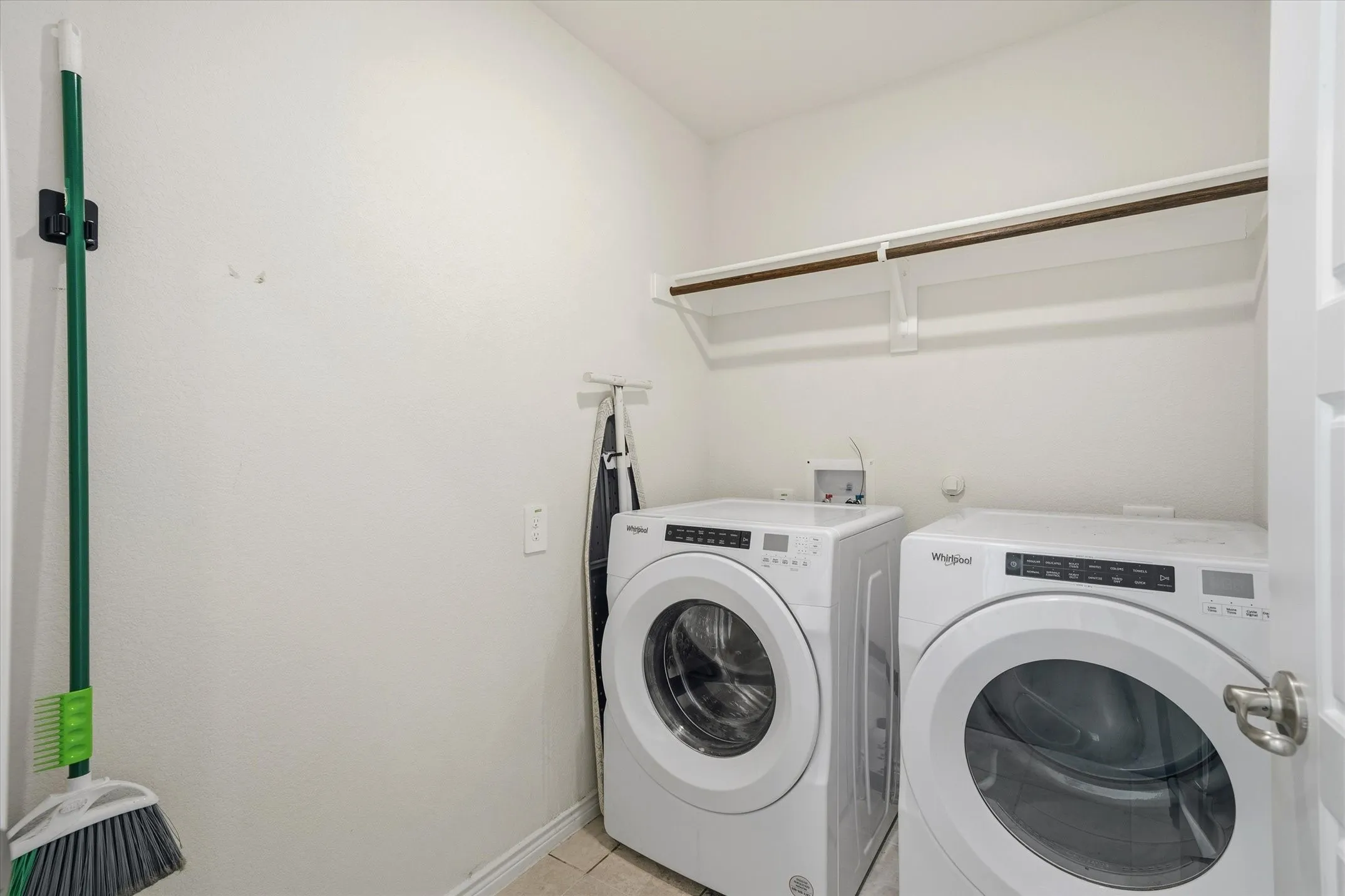 A dedicated interior utility room at 202 Buckboard Ln provides efficient laundry functionality with neutral textured walls and durable ceramic tile flooring. The space is equipped with side-by-side white front-loading Whirlpool washer and dryer units beneath an overhead storage shelf with an integrated clothes hanging rod. Recessed plumbing connections, organized wall mounts for cleaning tools, and bright finishes make this practical laundry area a tidy, move-in ready convenience within the home.