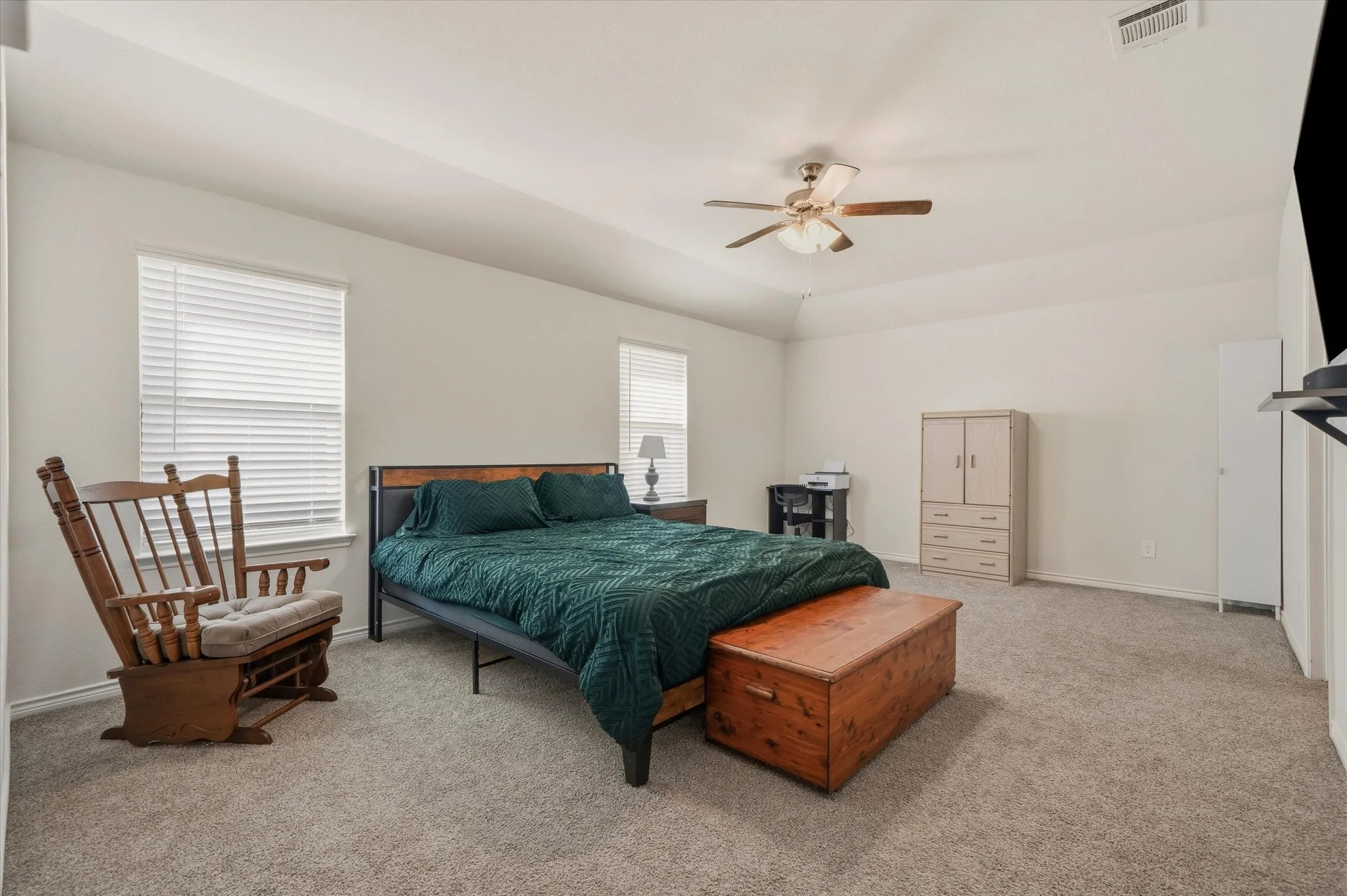 A spacious bedroom interior at 202 Buckboard Ln showcases high tray ceilings, neutral off-white walls, and plush beige carpeting. Twin single-hung windows equipped with horizontal blinds provide ample natural lighting across the room, while a central five-blade wood ceiling fan circulates air efficiently. The generous floor plan accommodates a king-size bed frame, accent glider seating, a versatile work desk, and multiple freestanding armoires, delivering a restful and highly functional living retreat inside this comfortable Caddo Mills home.