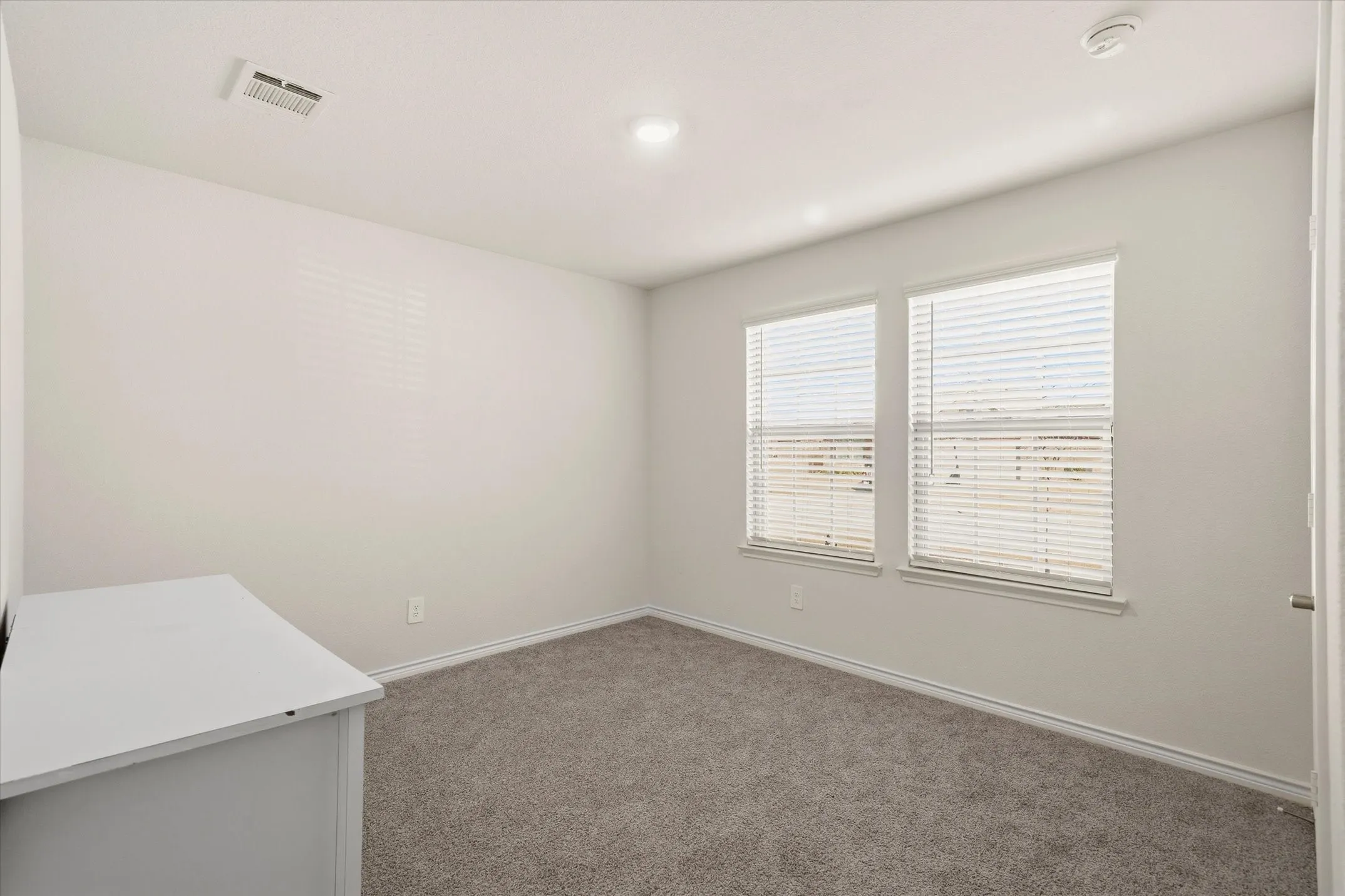 Natural light streams through dual windows in this modern bedroom at 202 Buckboard Ln in Caddo Mills. The interior showcases neutral walls, crisp white trim, plush carpeting, and recessed LED overhead lighting. Looking through the blinds reveals standard suburban lot setbacks, wooden privacy fencing, and views of neighboring single-story construction under clear Texas skies. This bright, adaptable space captures the quiet appeal of a newly developed residential neighborhood, offering comfortable everyday living within a growing master-planned community.