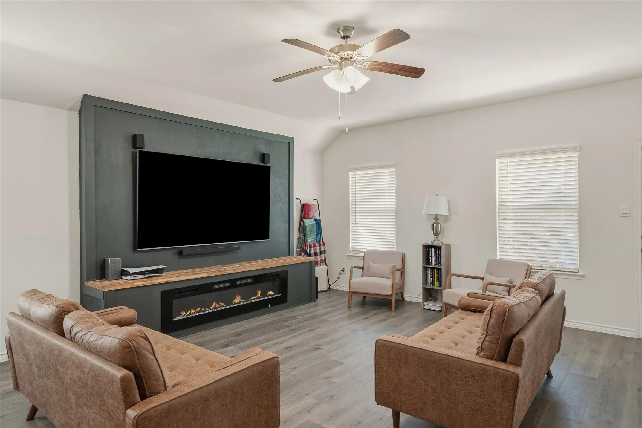 The living room at 202 Buckboard Ln presents a welcoming, open atmosphere defined by a dramatic vaulted ceiling and durable wide-plank, grey-toned wood-look flooring. A striking focal point anchors the space with a rich, dark matte green accent wall holding a mounted flat-screen television, natural wood mantle, and linear electric fireplace. Soft sunlight filters through two side windows dressed in horizontal blinds, illuminating the balanced seating arrangement and overhead brushed-nickel ceiling fan to create a stylish, functional everyday retreat in this Caddo Mills home.