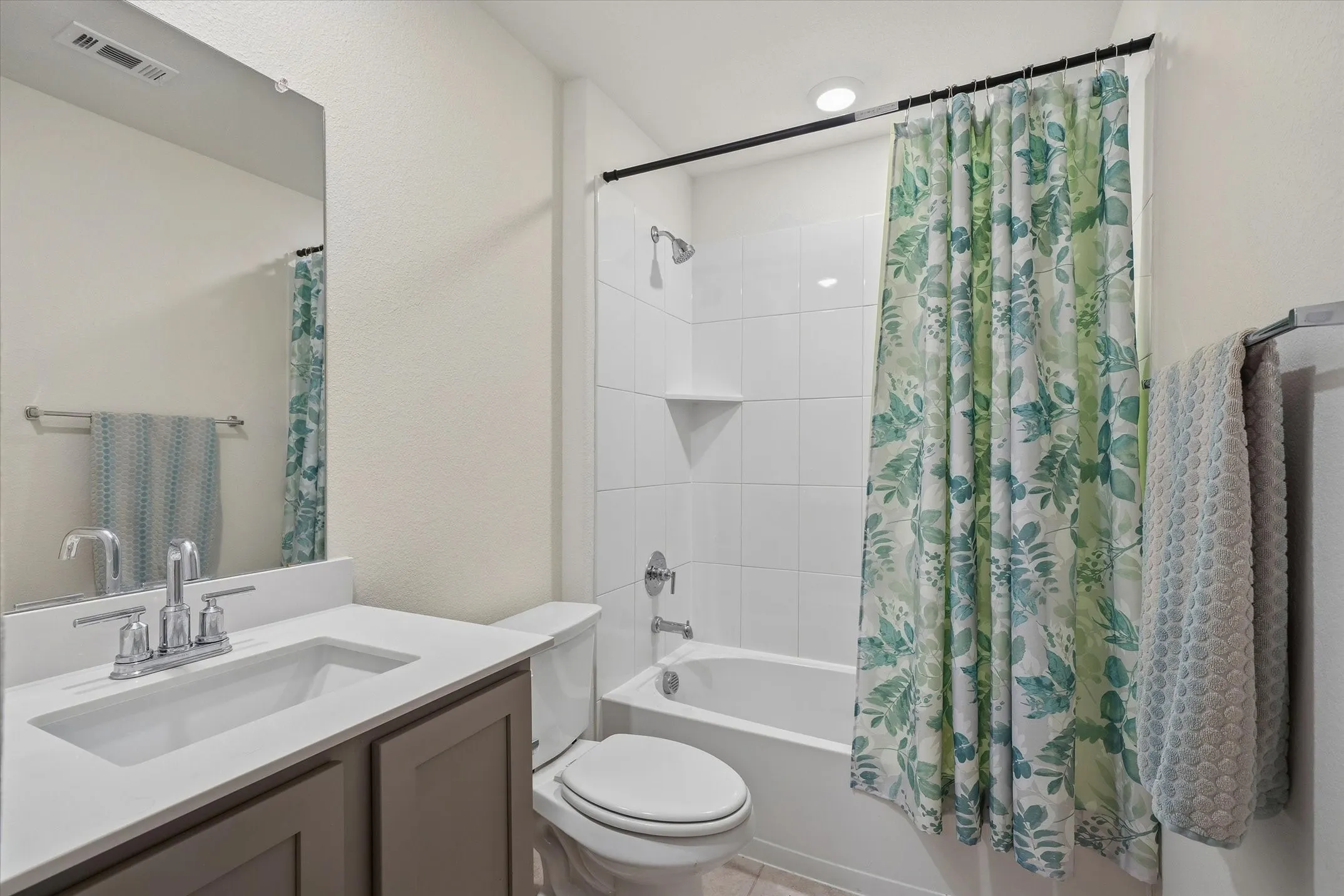 This full bathroom inside 202 Buckboard Ln presents a clean, functional layout highlighted by light tile flooring and neutral walls. A single-sink vanity features a dark taupe shaker-style base, solid white countertop, and a tall frameless mirror. Adjacent to the toilet, the built-in alcove bathtub and shower combination is lined with stacked white square tile reaching toward the ceiling, accented by sleek chrome fixtures and an overhead recessed light, creating a bright and pristine interior space for daily convenience.