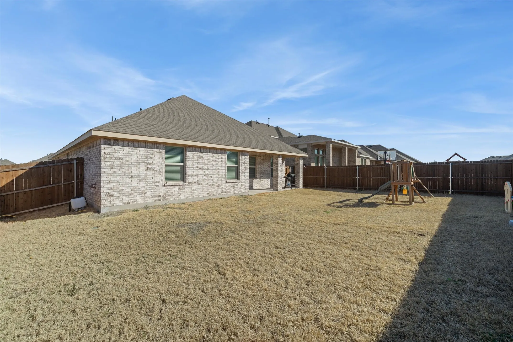 A wide view captures the expansive, fully fenced backyard of the single-story home at 202 Buckboard Ln. Flat, level terrain stretches across the rectangular lawn, completely enclosed by a tall wooden privacy fence with sturdy metal posts. The rear exterior showcases light variegated brick, a covered back patio, and a poured concrete slab foundation beneath a low-pitch hip roof. Mid-afternoon sun illuminates the open outdoor space, which easily accommodates a detached wooden playset and swing set while leaving ample room for outdoor recreation and custom landscaping.