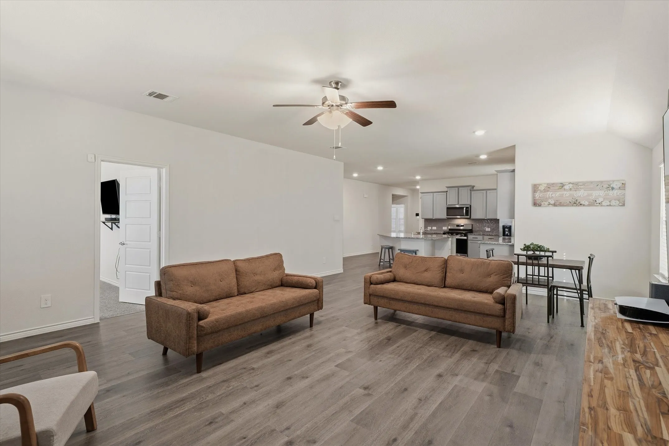 This bright, open-concept interior at 202 Buckboard Ln showcases a seamlessly connected living, dining, and kitchen layout ideal for modern entertaining. Neutral walls complement continuous grey wood-grain luxury vinyl plank flooring, recessed lighting, and a brushed-nickel ceiling fan. The staged gathering room transitions effortlessly toward an intimate dining alcove and a well-appointed kitchen featuring grey shaker cabinetry, granite-look countertops, an eat-in peninsula, and stainless steel appliances. An adjacent doorway reveals a secondary carpeted living space, accentuating the flexible design of this inviting Caddo Mills home.