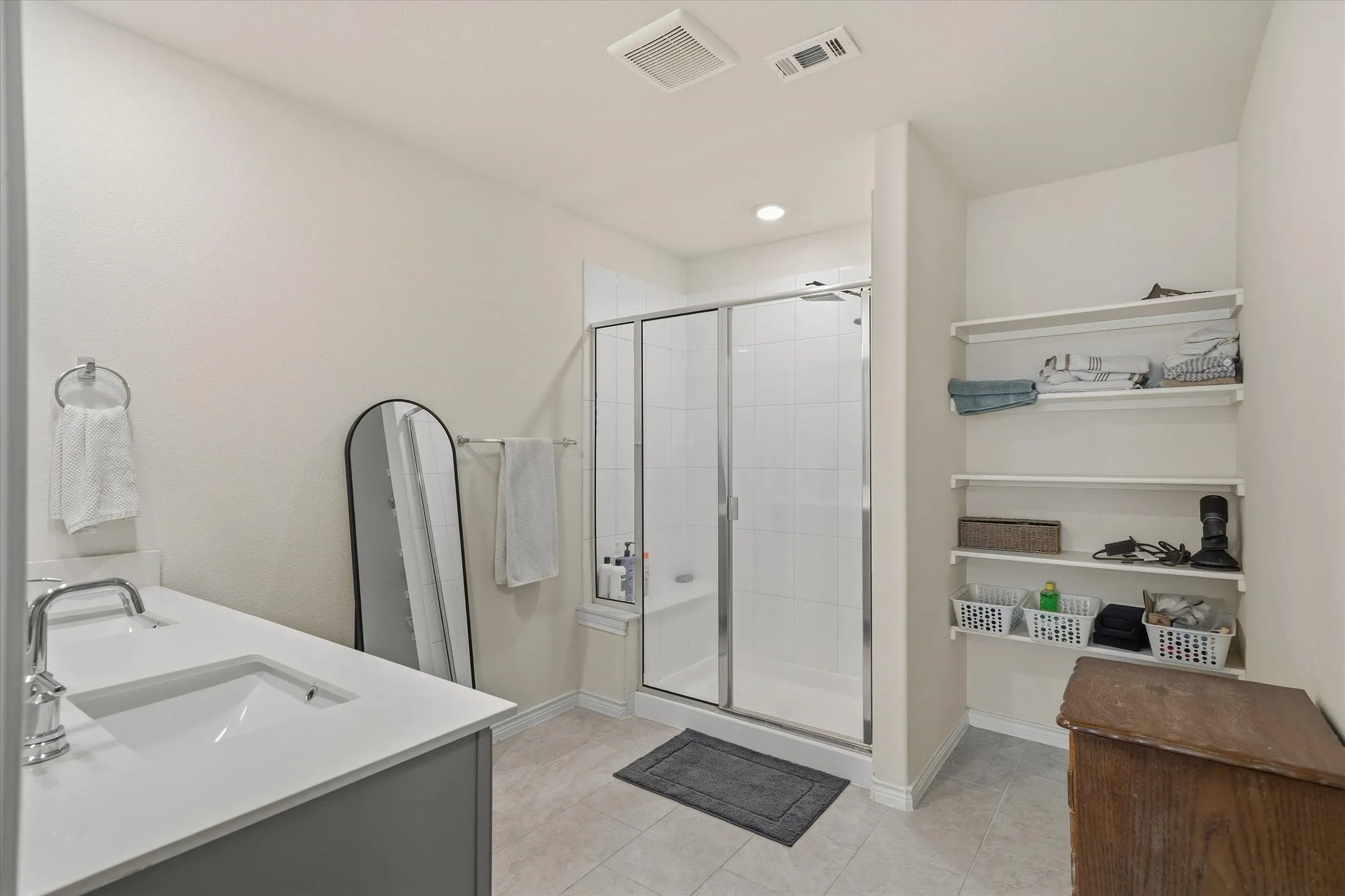 Primary bathroom interior at 202 Buckboard Ln, Caddo Mills, showcasing neutral ceramic tile flooring and light gray cabinetry with a crisp white solid-surface countertop. The functional layout features a contemporary undermount sink with chrome fixtures, an arched black-framed floor mirror, and a sliding glass-enclosed walk-in shower detailed with white wall tile and a built-in bench. An organized linen alcove with open shelving provides dedicated storage alongside clean drywall finishes, offering a bright, move-in-ready ensuite retreat.