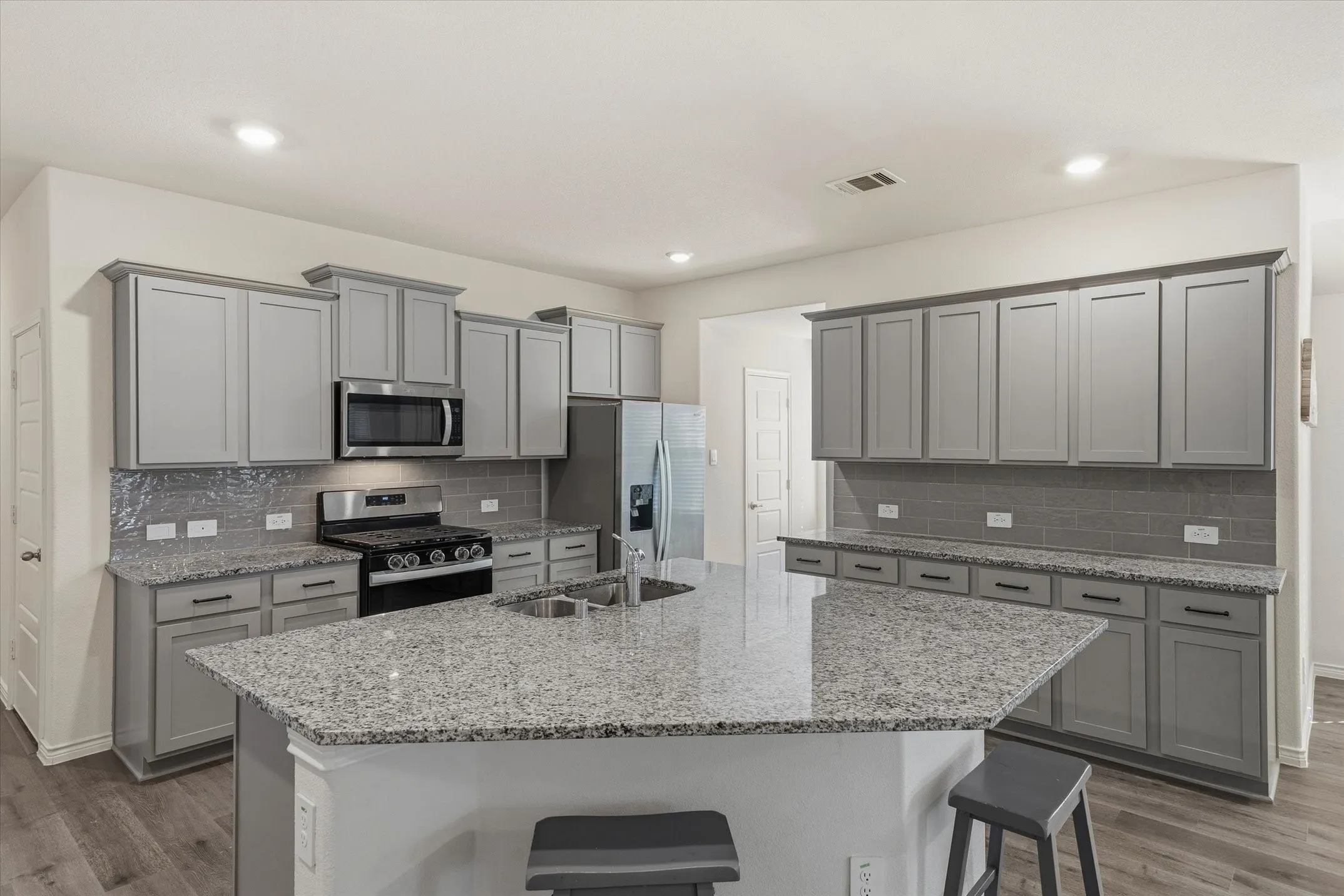 Inside 202 Buckboard Ln, this open-concept kitchen features an expansive angled island fitted with speckled granite countertops, an undermount double-basin sink, and breakfast-bar seating. Sleek light-gray shaker cabinetry with dark hardware coordinates with a gray subway tile backsplash and durable light wood-look plank floors. Quality stainless-steel appliances include a gas range, mounted microwave, and French-door refrigerator, all illuminated by recessed LED ceiling lights. The thoughtful layout provides seamless flow to adjoining living areas, delivering an ideal culinary centerpiece for modern Texas living.