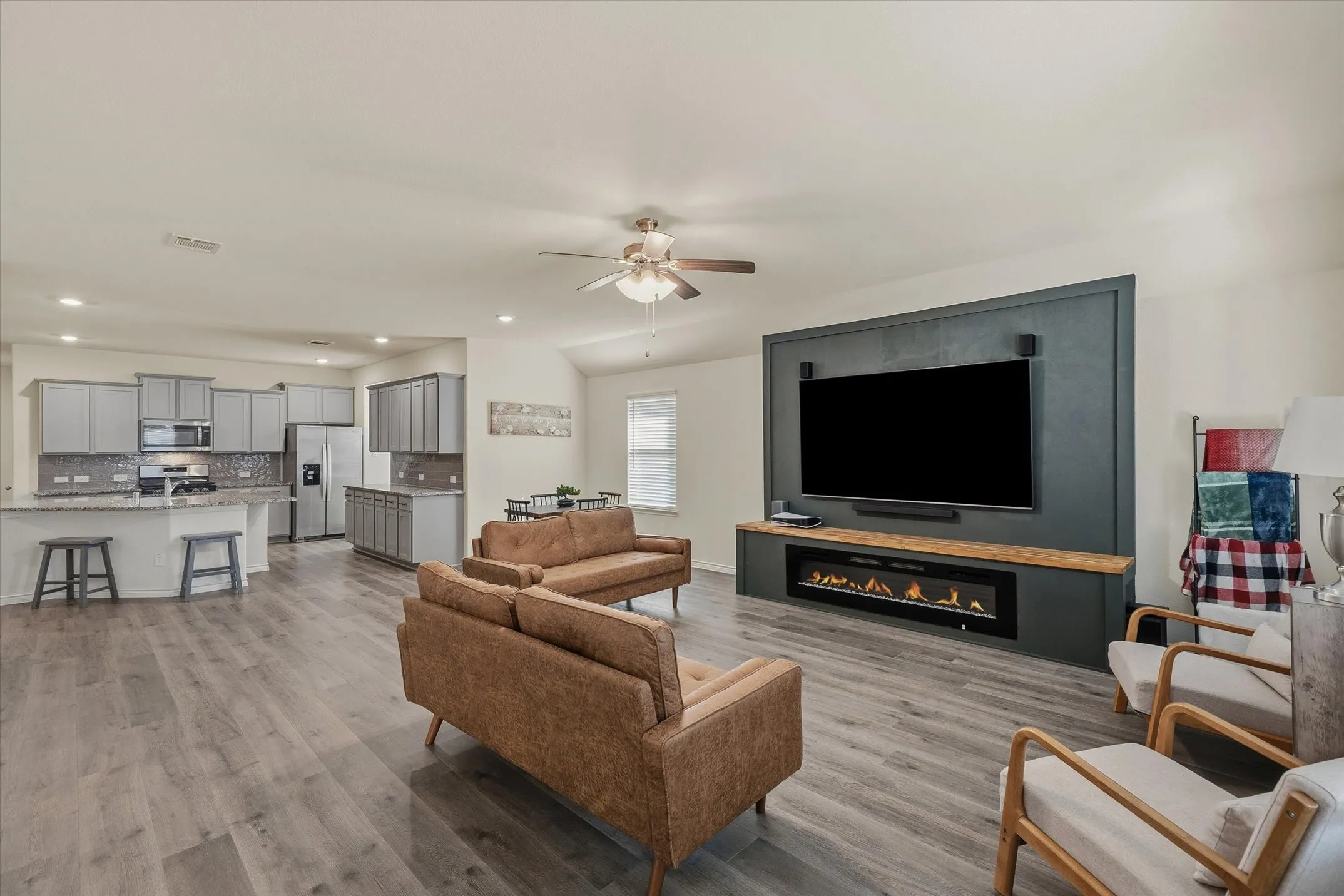 The bright open-concept living space at 202 Buckboard Ln features modern wood-look laminate flooring that seamlessly connects the family room, dining nook, and kitchen. A striking dark accent media wall with a built-in linear electric fireplace anchors the living area beneath recessed LED lighting and a contemporary ceiling fan. In the background, the functional kitchen showcases painted shaker cabinets, granite countertops, and stainless steel appliances, creating an inviting layout ideal for relaxed everyday living and effortless indoor entertaining in Caddo Mills.