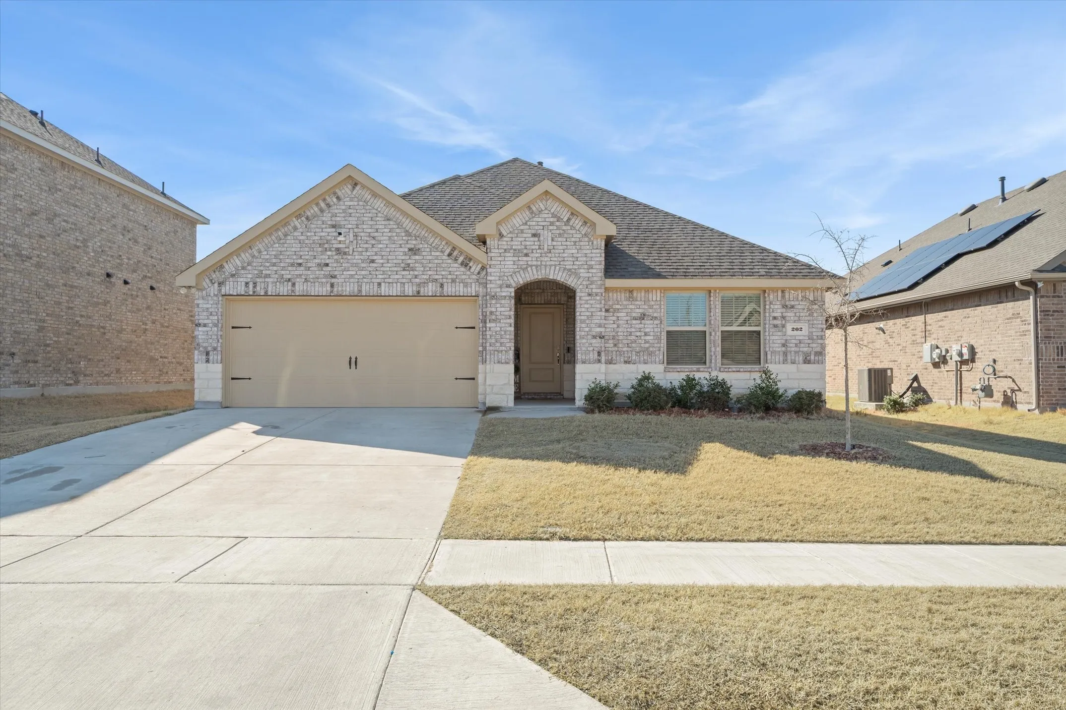 This single-story residence at 202 Buckboard Ln features charming curb appeal within a quiet suburban neighborhood. The home showcases light-toned brick masonry, a neat limestone water table accent, and a multi-gabled dark shingle roofline. A concrete driveway leads to an attached two-car garage with carriage-style hardware, alongside an arched, sheltered front entryway. The level front lot includes a concrete sidewalk, low-profile foundation plantings, and a young shade tree, providing an inviting exterior finish under open Texas skies.