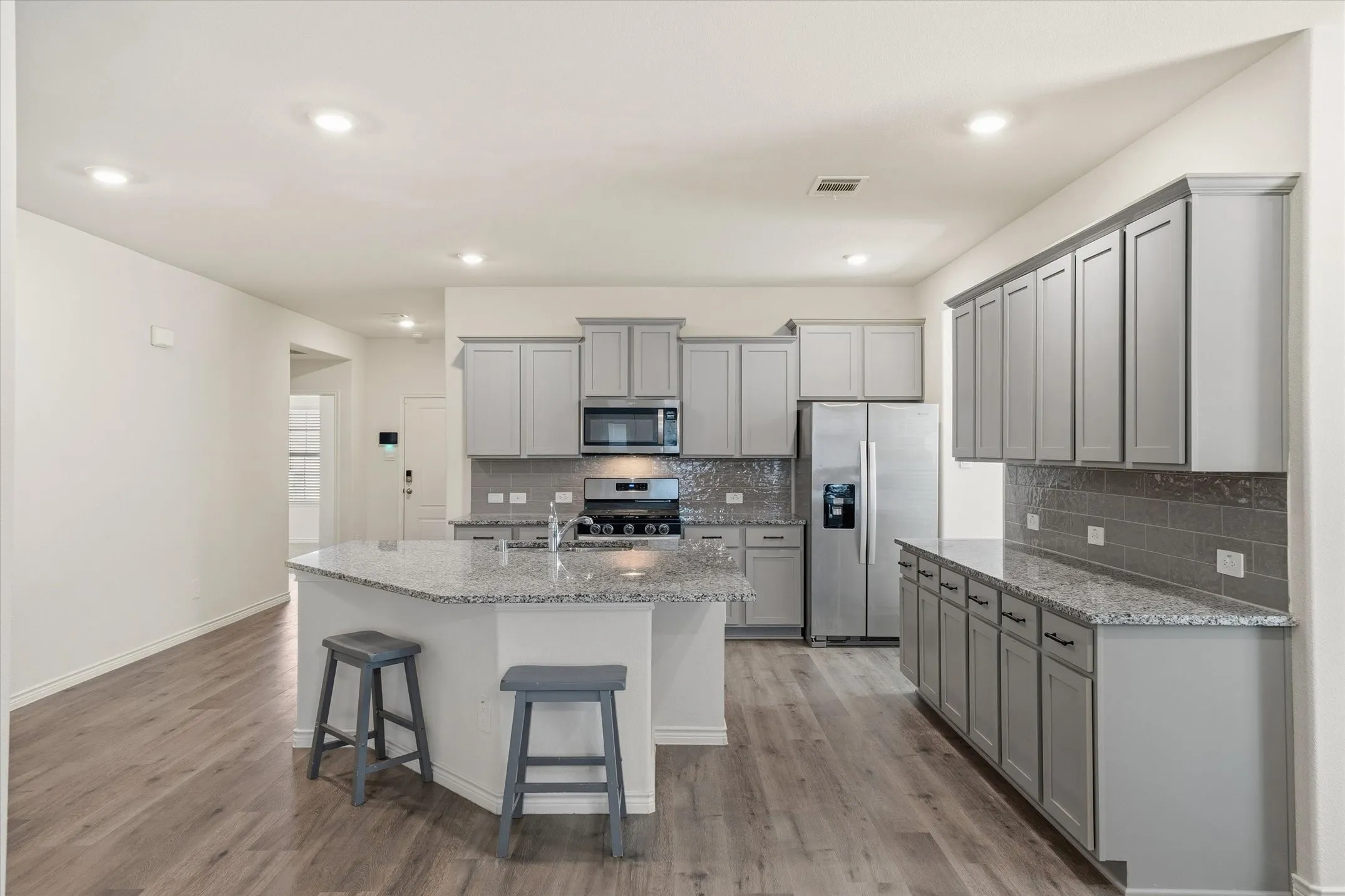 Inside 202 Buckboard Ln, this contemporary kitchen features a seamless open-concept design complete with durable grey wood-look plank flooring. The space is anchored by an angled prep island with breakfast bar seating and an integrated sink, highlighted by speckled granite countertops and glossy subway tile backsplashes. Stainless steel appliances, including a gas range and side-by-side refrigerator, complement the light grey Shaker cabinetry with modern black hardware. Recessed ceiling lights illuminate the inviting layout, providing smooth sightlines into the adjoining living spaces of this Caddo Mills residence.