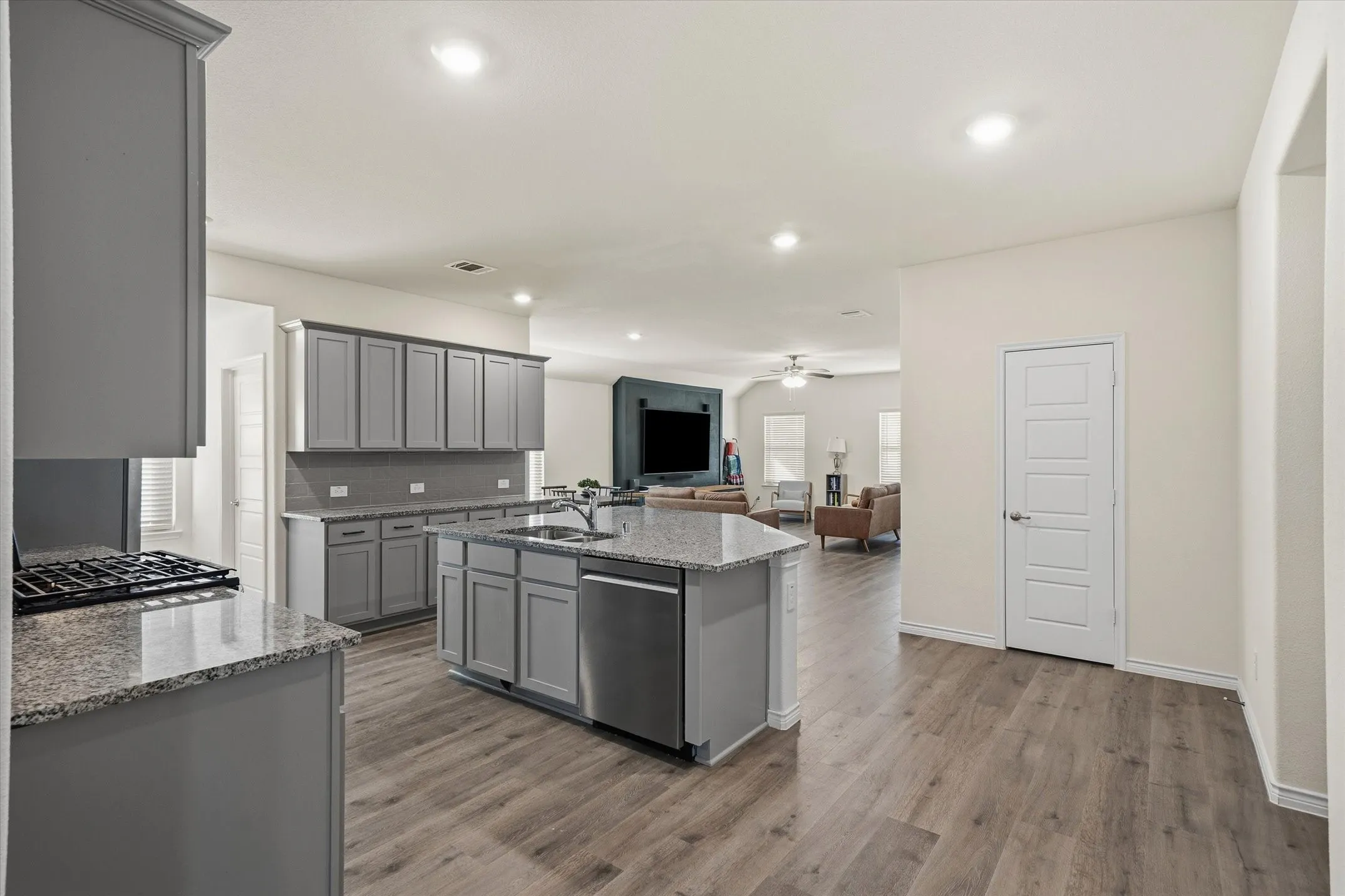 This open-concept interior at 202 Buckboard Ln showcases seamless suburban living, connecting a modern kitchen to the primary living space. Light grey shaker cabinetry, granite countertops, and a subway-tile backsplash frame the culinary area, centered by an expansive prep island with an undermount double sink and breakfast bar seating. Continuous wood-look luxury vinyl plank flooring unites the entire main level beneath recessed lighting, leading directly to a welcoming living room complete with built-in cabinetry, an entertainment wall, and bright windows fitted with horizontal blinds.