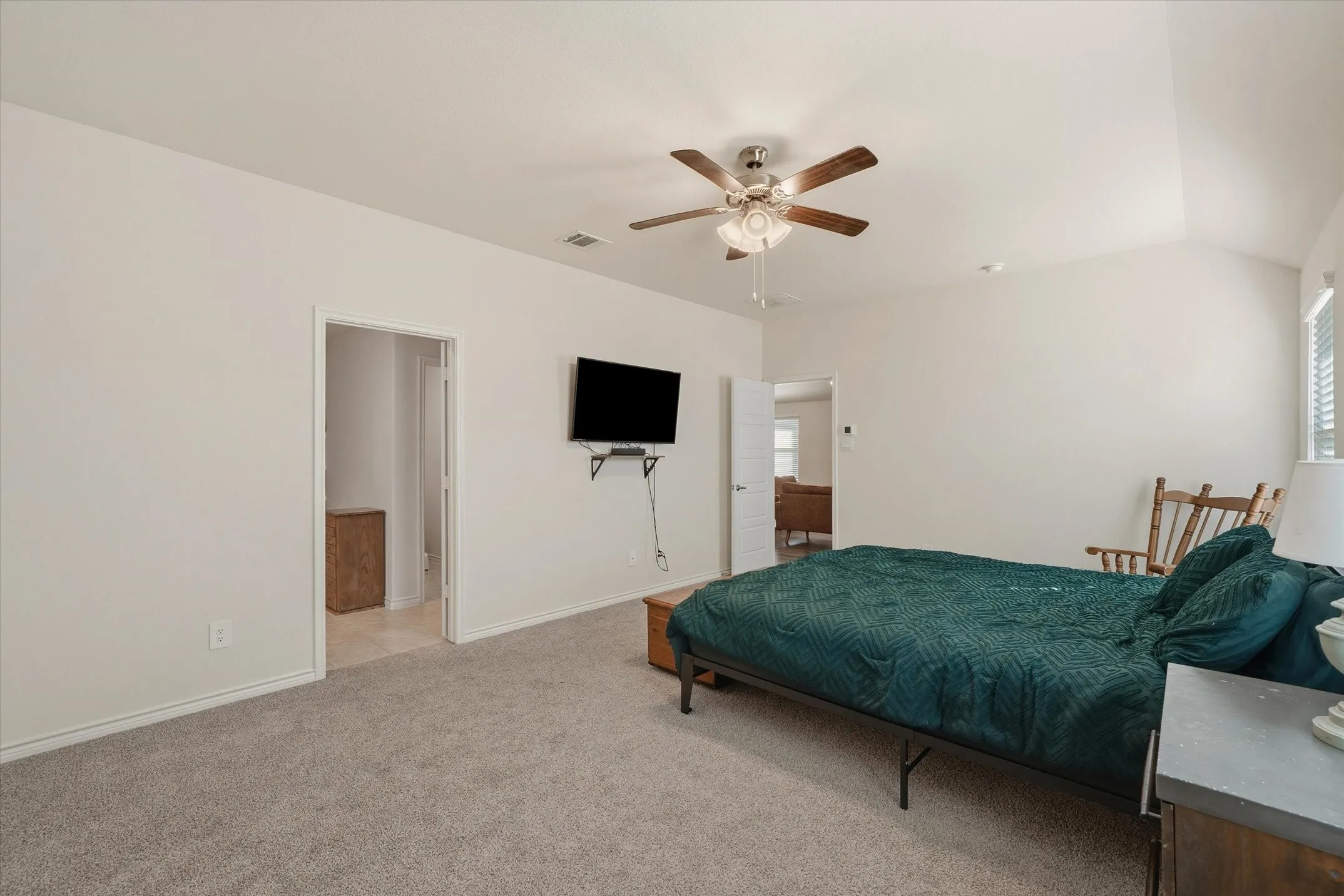 A spacious primary bedroom suite at 202 Buckboard Ln offers a restful retreat featuring plush neutral carpeting, soft beige walls, and high ceilings with an integrated ceiling fan. Natural light pours in through side windows fitted with horizontal blinds, illuminating room for a king-size bed and sitting furniture. The functional layout provides immediate access to an attached en-suite bathroom with tiled floors as well as an open doorway leading back to the home's main living areas, delivering both everyday convenience and privacy.