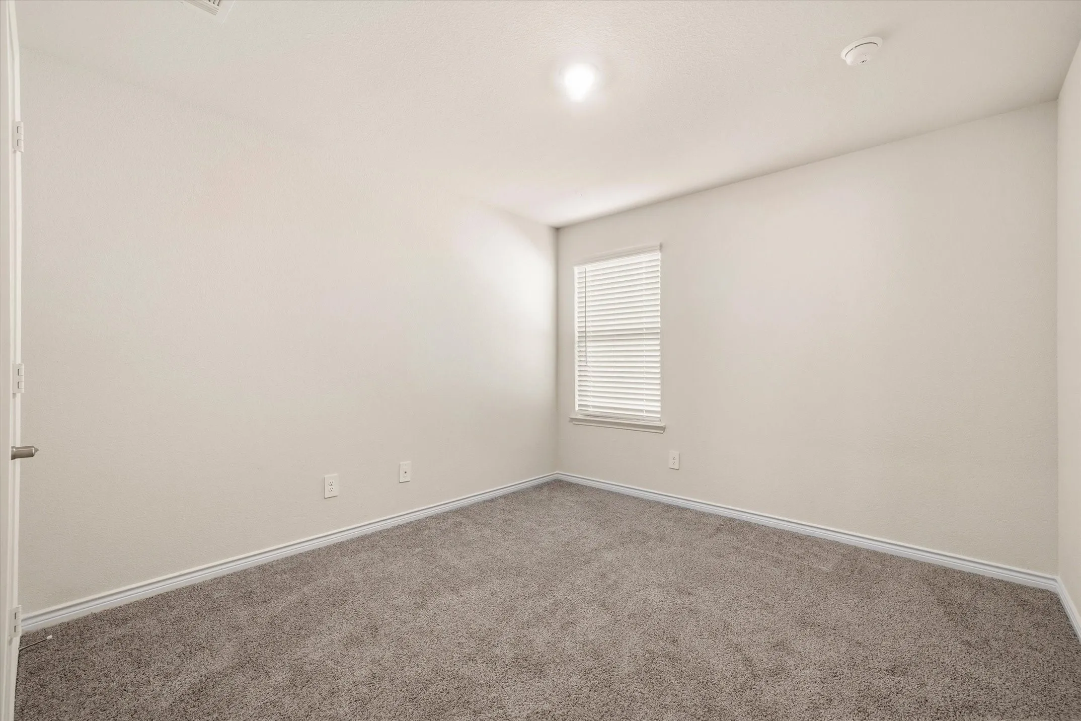 Inside this move-in-ready residence at 202 Buckboard Ln in Caddo Mills, a secondary bedroom presents a versatile, well-lit retreat. The functional interior showcases soft, neutral-toned walls complemented by clean white baseboards and plush, heathered wall-to-wall carpeting underfoot. Natural daylight streams through a tall exterior window fitted with horizontal blinds, balanced by central overhead lighting and dedicated HVAC venting. Complete with modern electrical outlets and a standard entry door, this bright space easily adapts to a private guest suite, home office, or quiet personal haven.