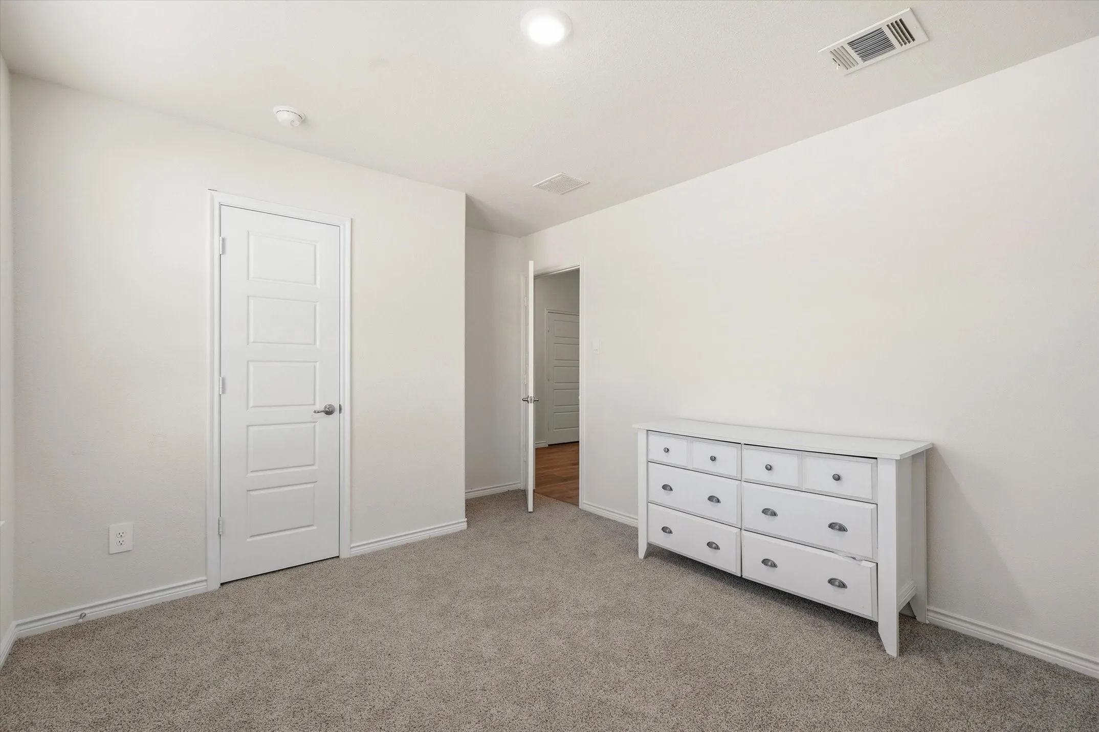 Interior view of a pristine secondary bedroom at 202 Buckboard Ln, highlighting clean architectural finishes and move-in-ready appeal. The space features plush neutral carpeting, fresh light-beige walls, crisp white trim, and stylish five-panel doors fitted with brushed nickel hardware. Overhead, recessed LED lighting and flush-mounted HVAC vents ensure optimal comfort throughout. An open doorway provides a seamless visual transition to the adjacent hallway with wood-look plank flooring, offering a clear perspective on this Caddo Mills residence layout and construction quality.