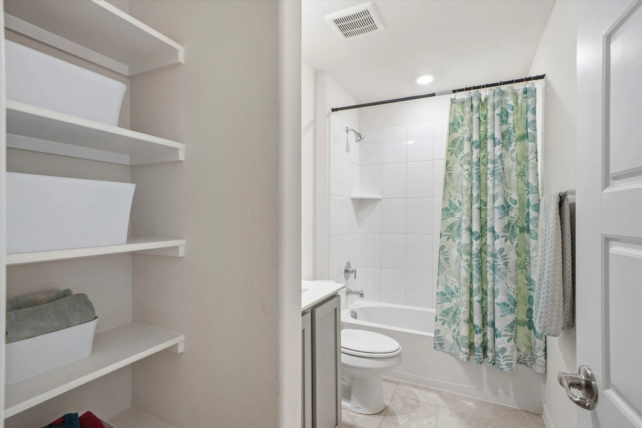 Inside 202 Buckboard Ln in Caddo Mills, this well-appointed full bathroom showcases practical organization and neutral finishes. A deep built-in linen alcove features tiered floating shelves for convenient towel and toiletry storage. Clean ceramic tile flooring extends across the room toward the single-sink vanity, commode, and full bathtub-shower combination lined with classic white ceramic wall tile and chrome fixtures. Recessed ceiling lighting completes this bright, functional bath space designed for effortless daily living.