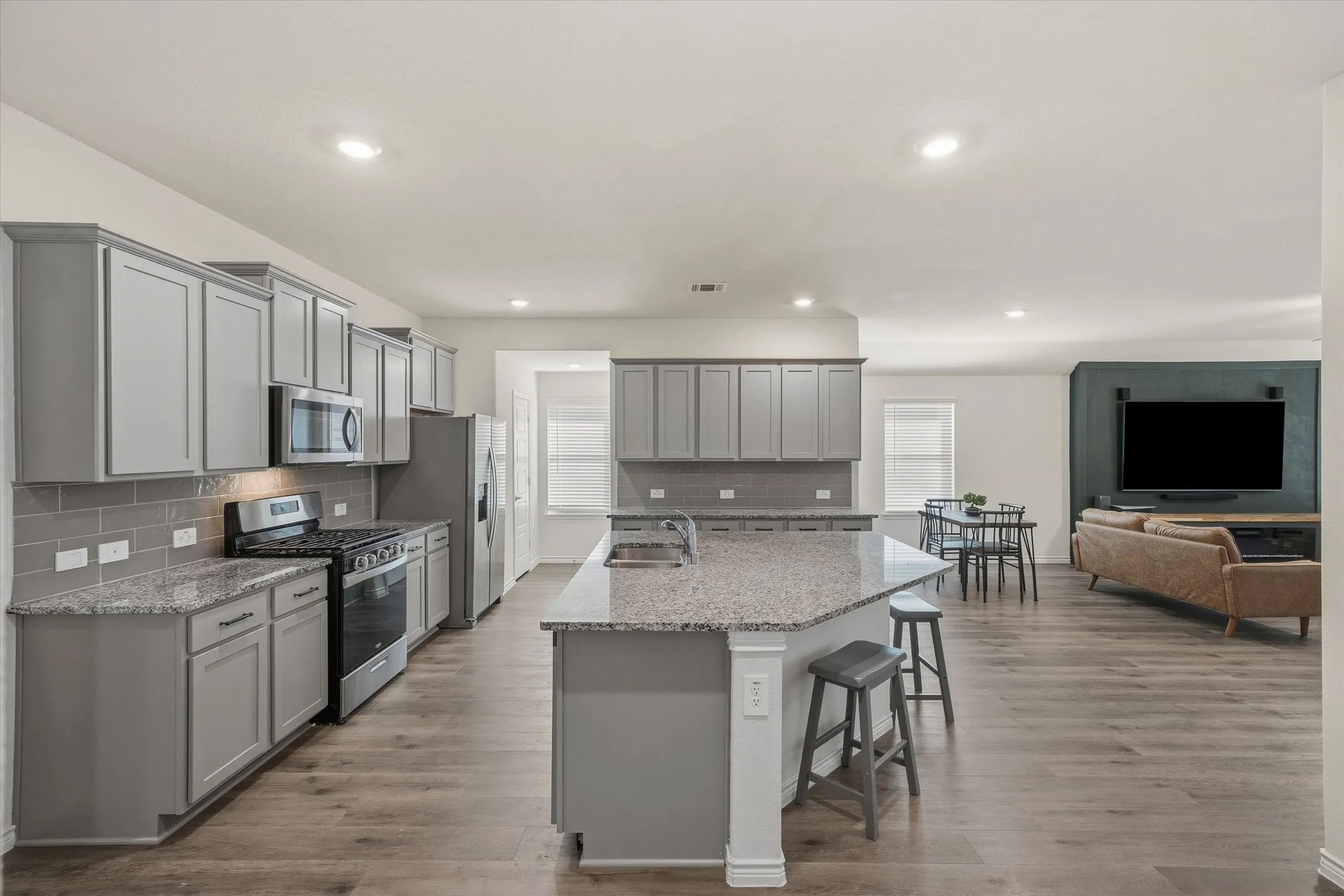 This open-concept interior at 202 Buckboard Ln highlights seamless entertaining with durable gray wood-look plank flooring connecting the kitchen, dining, and living zones. The modern kitchen boasts gray Shaker cabinetry, speckled granite countertops, a subway tile backsplash, stainless steel appliances, and an angled eat-in island with breakfast bar seating. Recessed lighting brightens the kitchen and adjacent dining area, which flows naturally into the spacious great room featuring a bold dark accent wall, creating a stylish, cohesive layout ideal for everyday modern living.