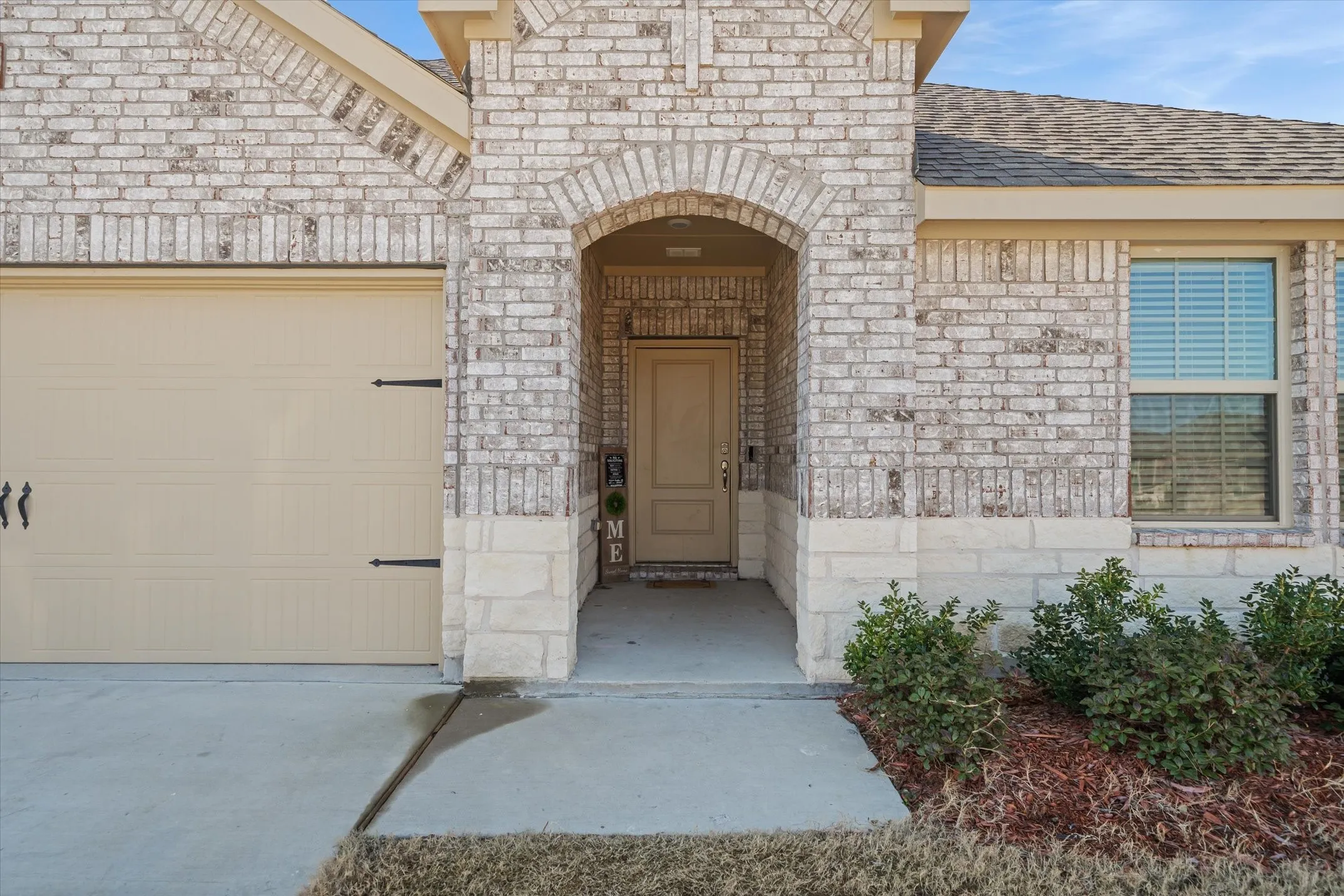 Eye-level view of the front exterior at 202 Buckboard Ln, highlighting a modern single-story residence crafted with whitewashed brick and rough-cut limestone wainscoting. A graceful brick arch shelters the recessed entryway, complemented by a warm tan front door and a matching sectional garage door with decorative carriage-house hardware. A poured-concrete driveway and front walkway cross the level lawn, bordered by tidy red-mulch landscaping beds with evergreen shrubs, all presented beneath a clear sky in Caddo Mills.