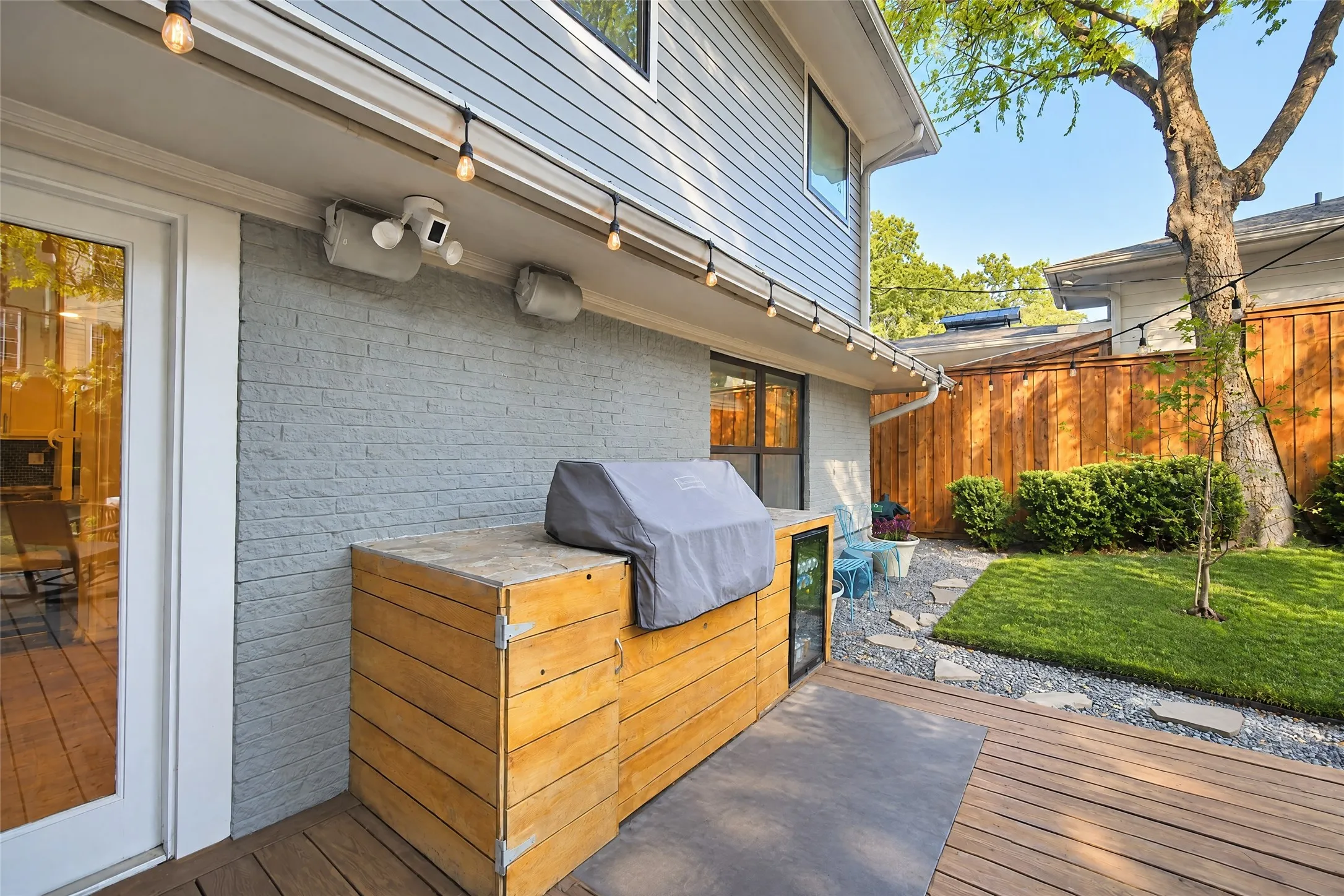 Expansive outdoor living area featuring a wood-finish deck, built-in outdoor kitchen, string lighting, and a manicured lawn area with stone pathway