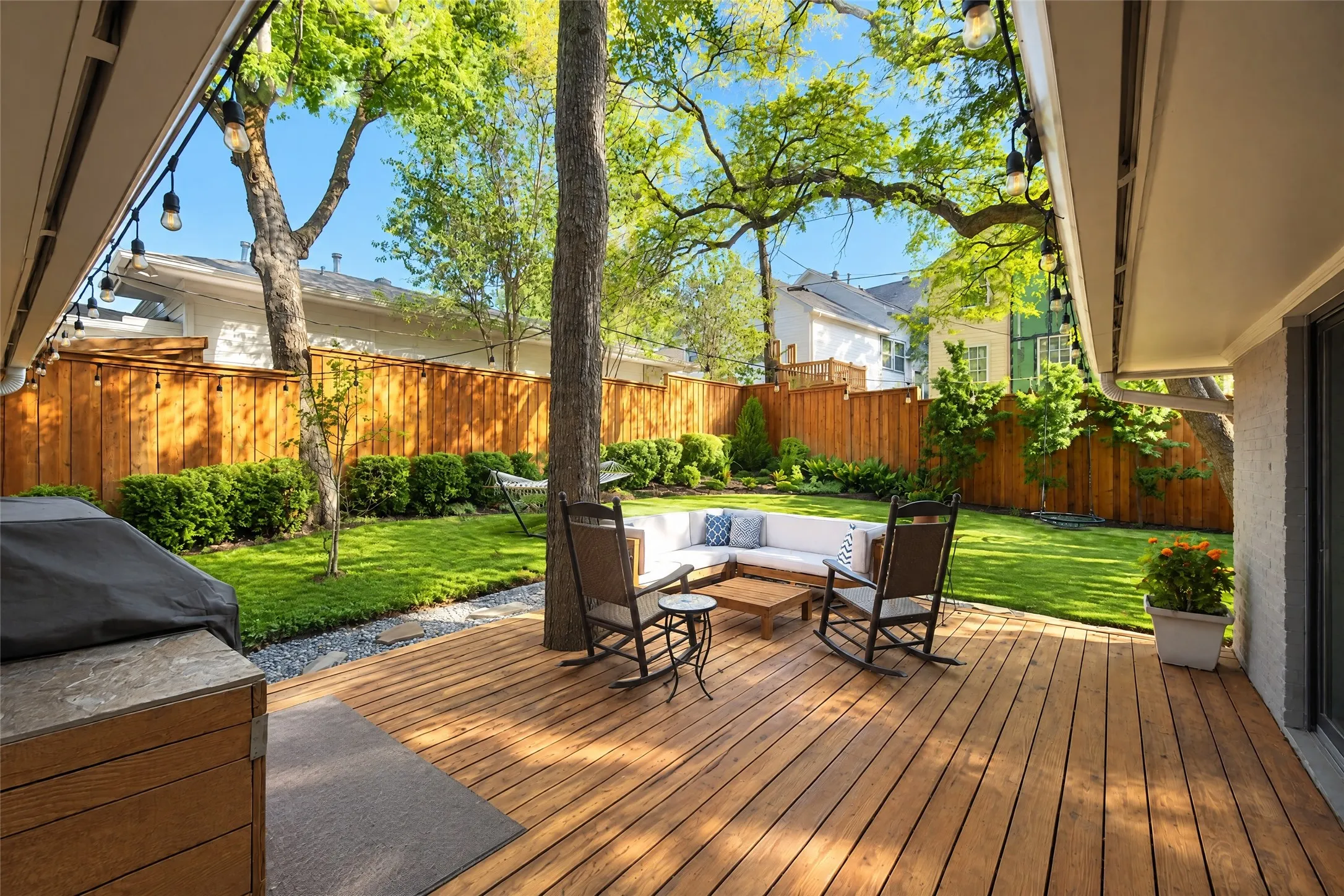 Expansive wooden deck, string lighting, manicured lawn, full privacy fencing, and mature trees