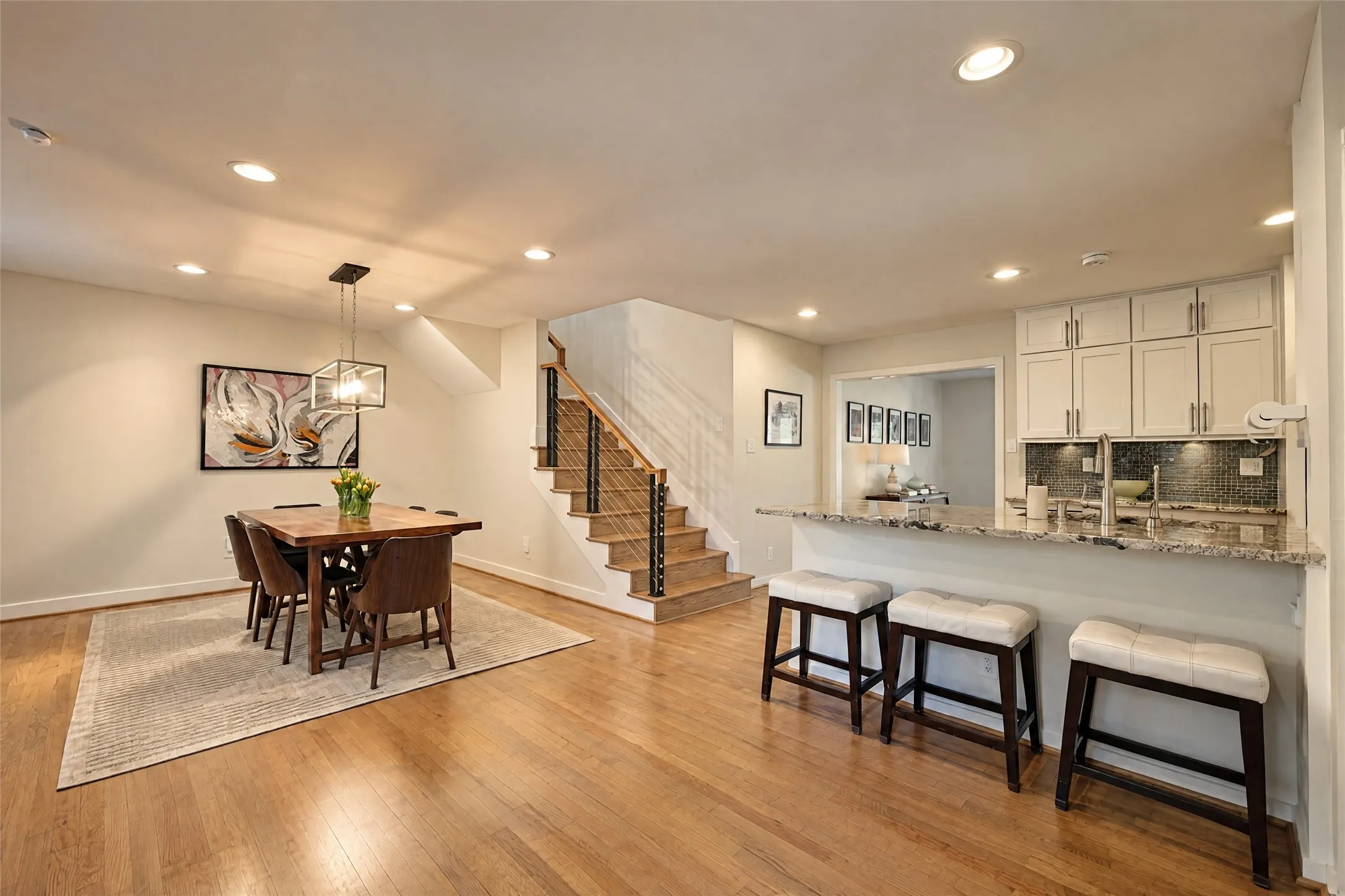 Hardwood flooring throughout, recessed lighting, modern stair railing with wood treads, kitchen island with stone countertop, and white kitchen cabinetry with backsplash