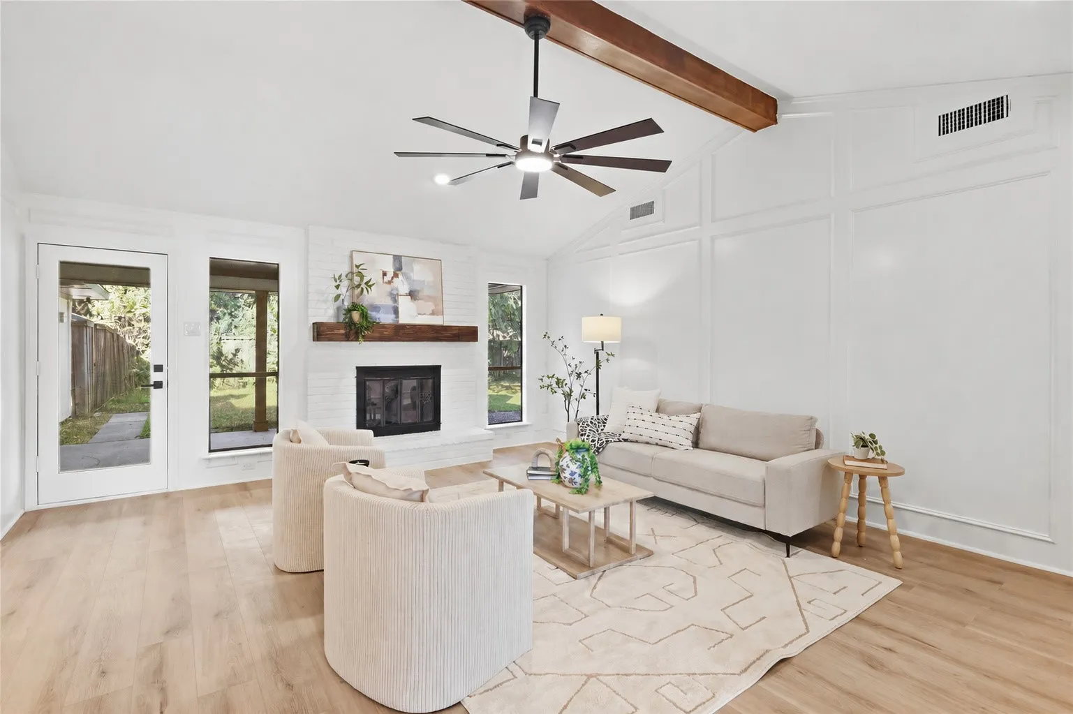 Vaulted ceiling with exposed wood beam, wood-finish flooring, white brick fireplace with wood mantel, paneling accent wall, and a ceiling fan with integrated lighting