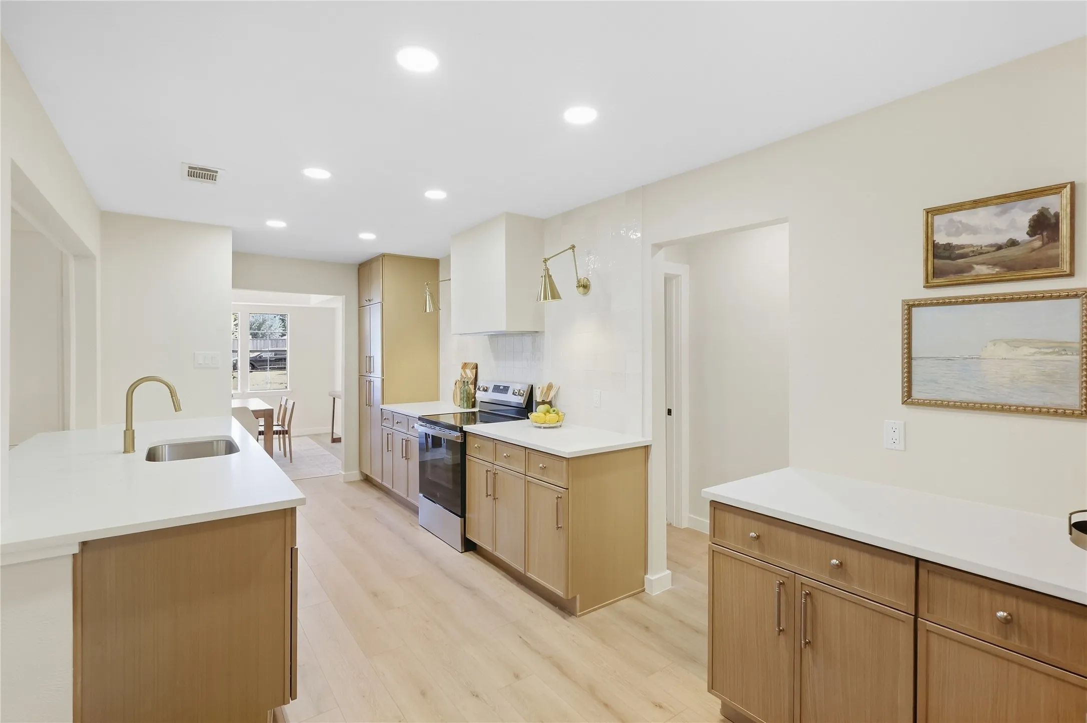 Recessed ceiling lighting illuminates the kitchen featuring wood-finish cabinetry, white countertops, a stainless steel sink with a gold-tone faucet, and a tile backsplash