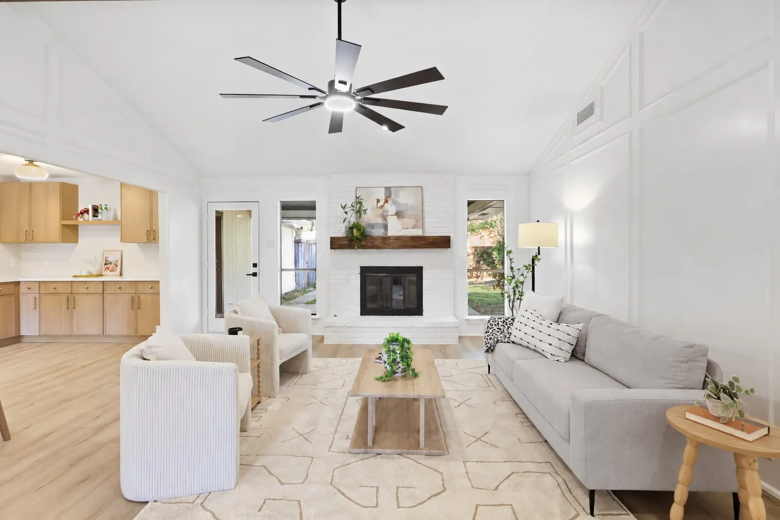 Vaulted ceiling with modern ceiling fan, brick fireplace with wood mantel, wood-finish flooring, white paneled wall accents, recessed lighting