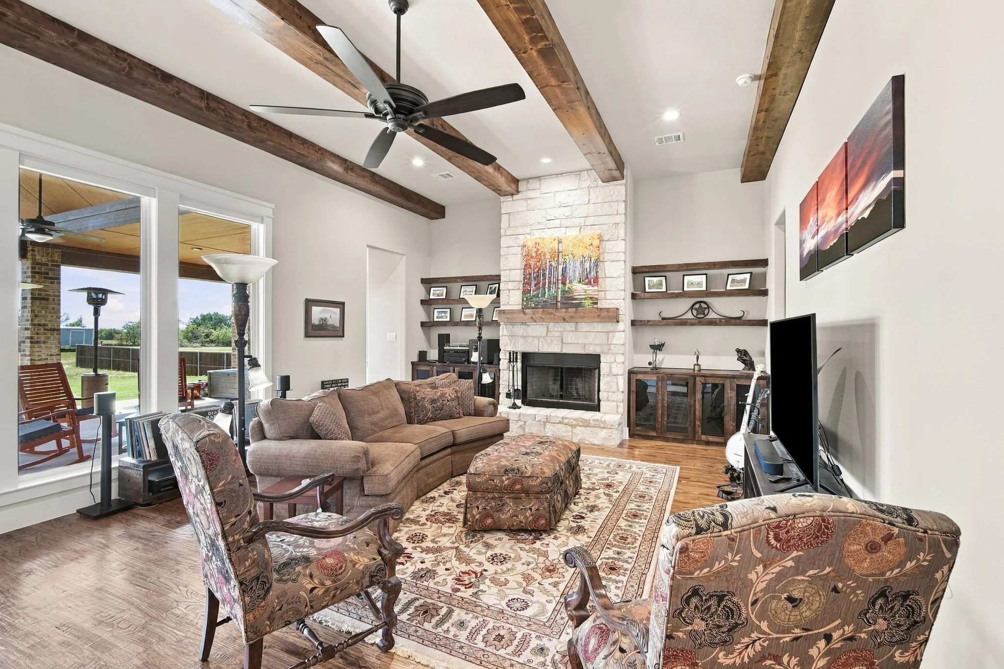 Living area features wood beams, a stone- fireplace with a hearth and wood mantle. Built-in cabinets with floating shelves offer a spot for special things.