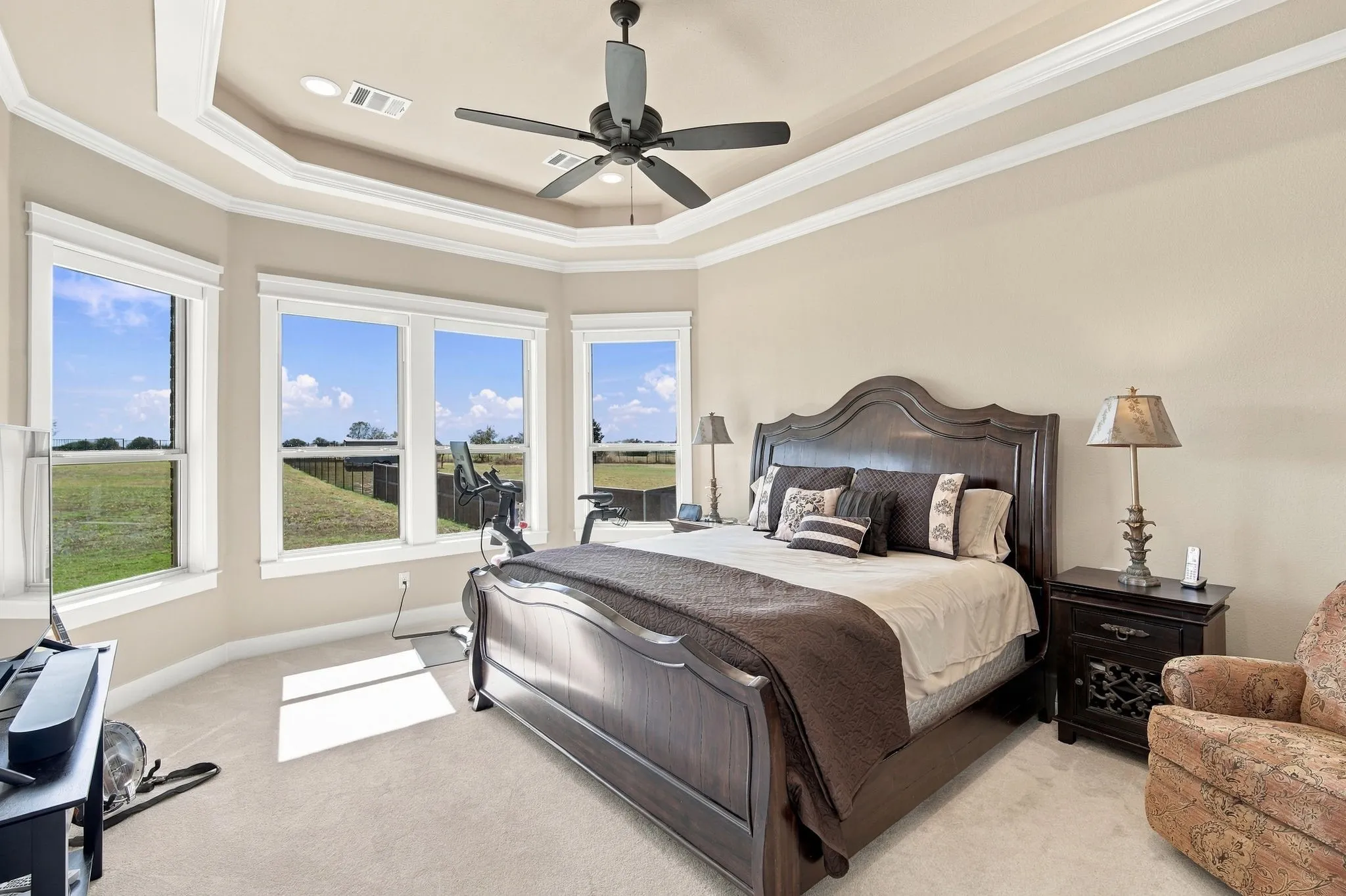 Elegant primary bedroom with a Tray ceiling with crown molding and cove lighting. Wood casing around the  windows and Electric shades.