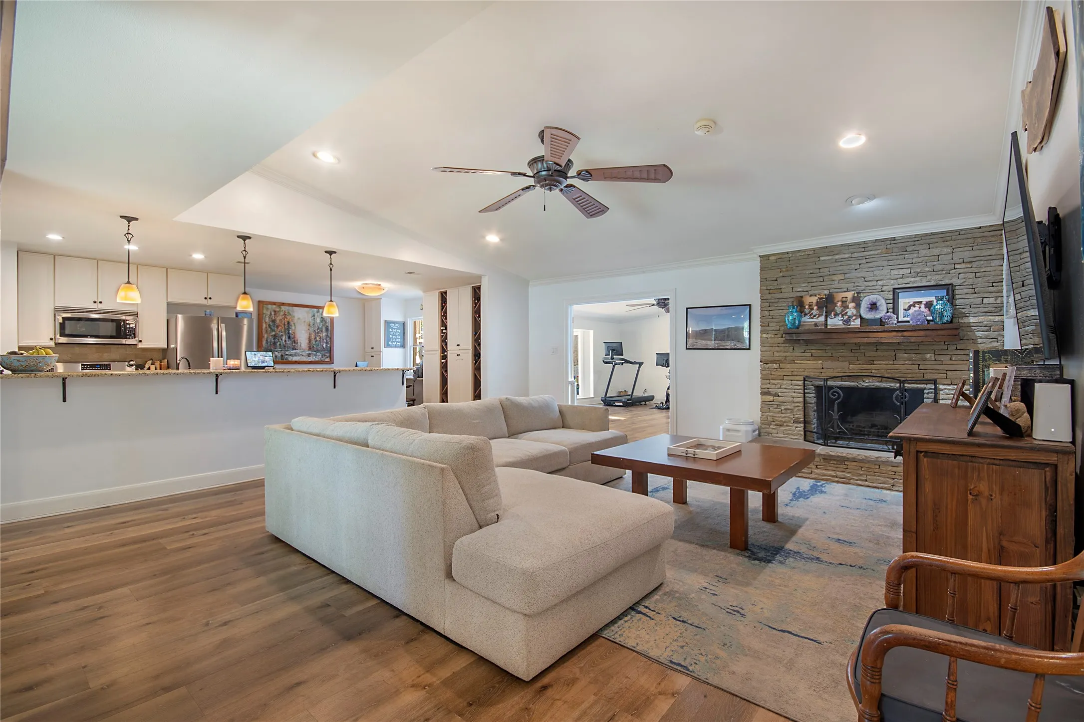Open-concept living space featuring wood-finish flooring, a stacked stone fireplace, a kitchen with white cabinetry, and stainless steel appliances