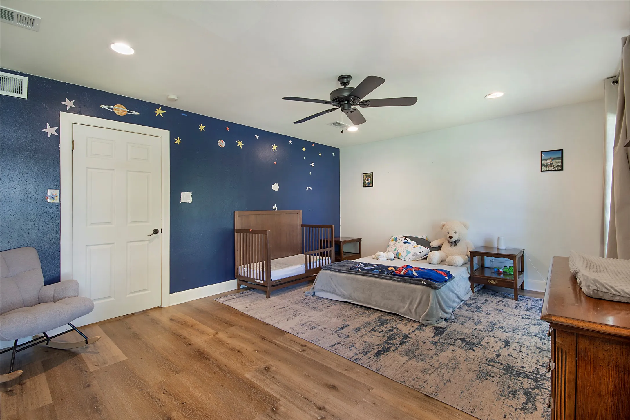 Spacious bedroom featuring warm wood-finish flooring, a ceiling fan with dark metal blades, recessed lighting, and a deep navy accent wall adorned with decorative celestial details
