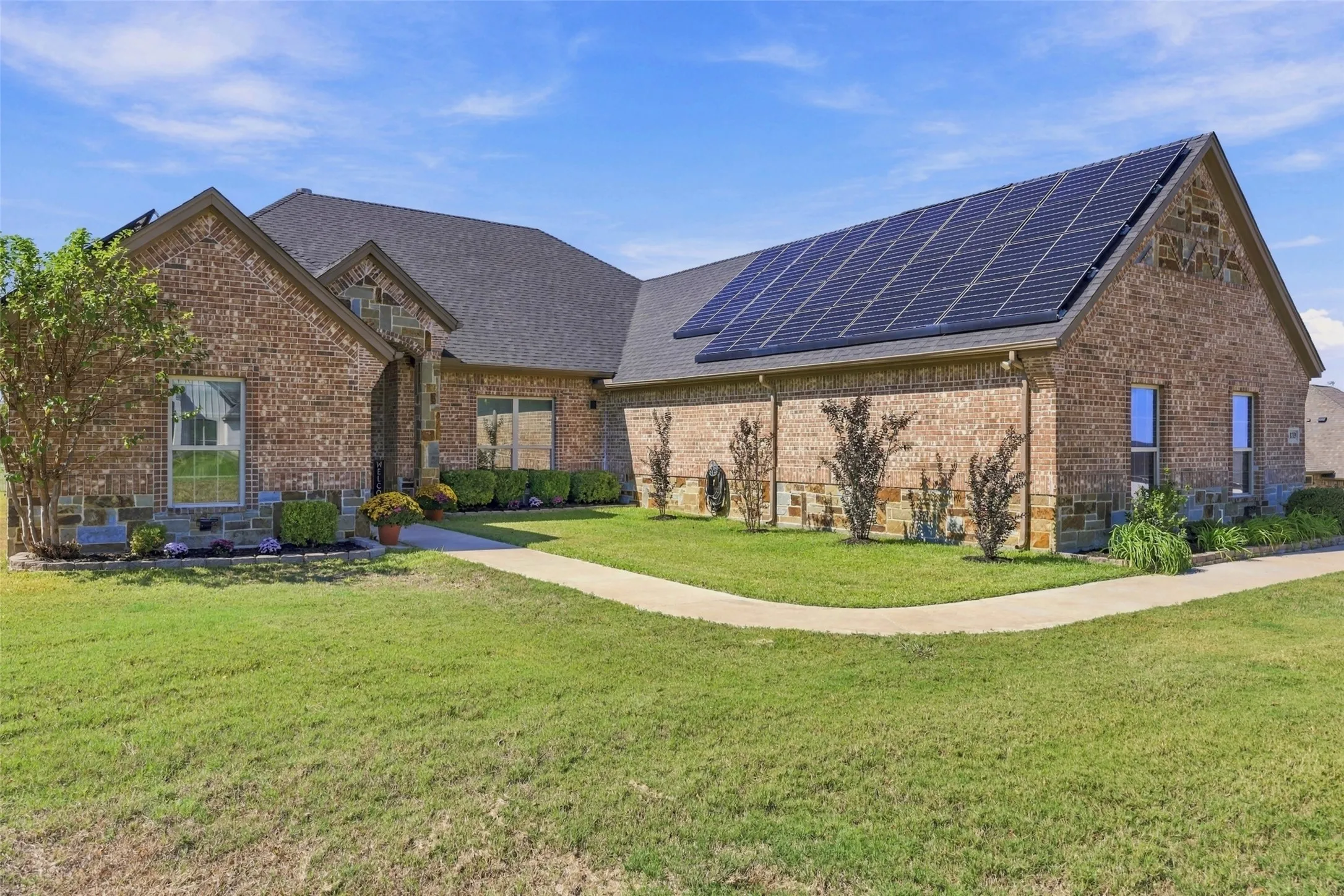 Brick exterior with stone accents, dark shingle roofing, multiple solar panels, and a concrete walkway